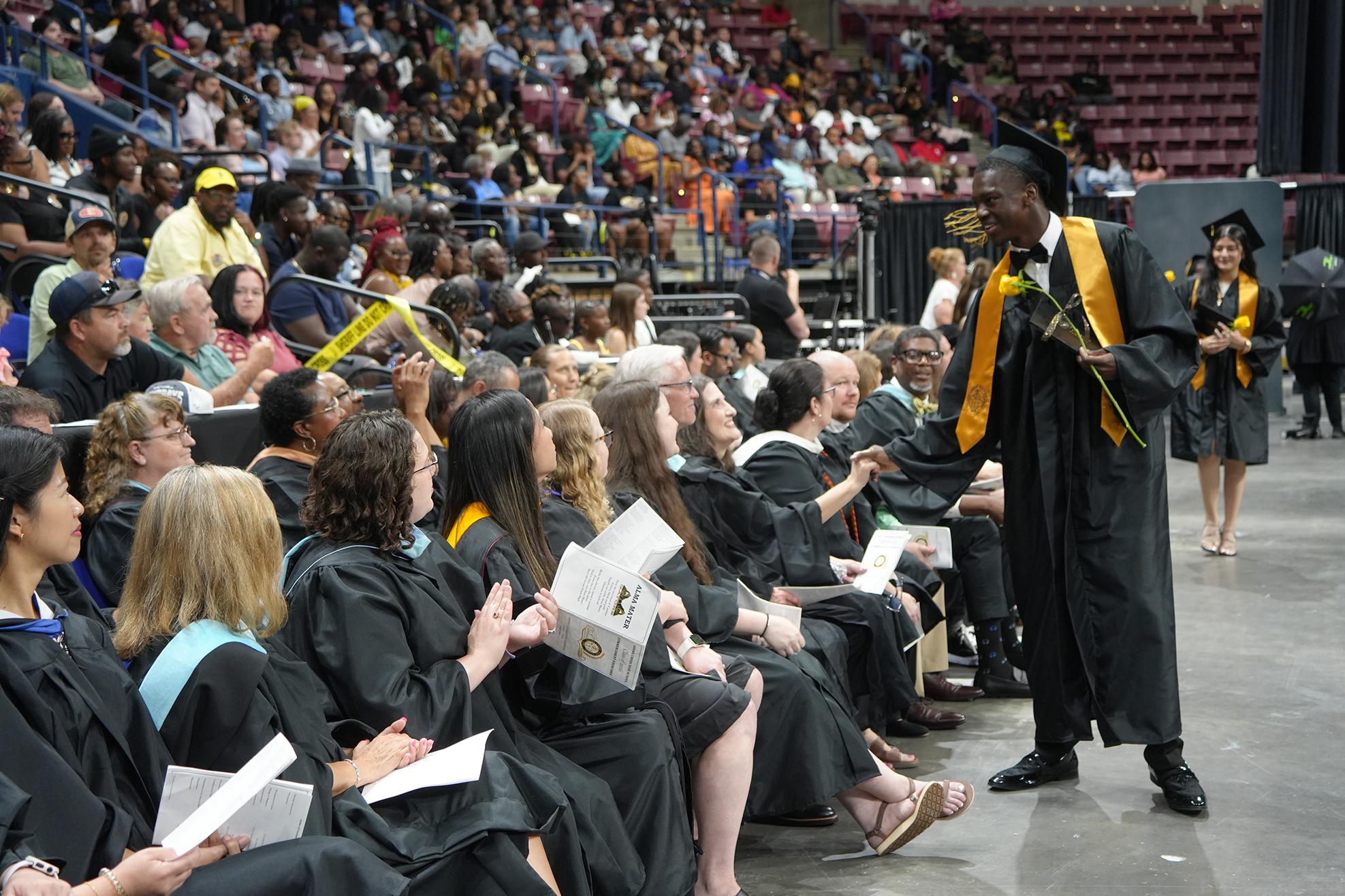 A man in a black cap and gown holds a rose while standing on a stage. Many people in black gowns sit in front of him.