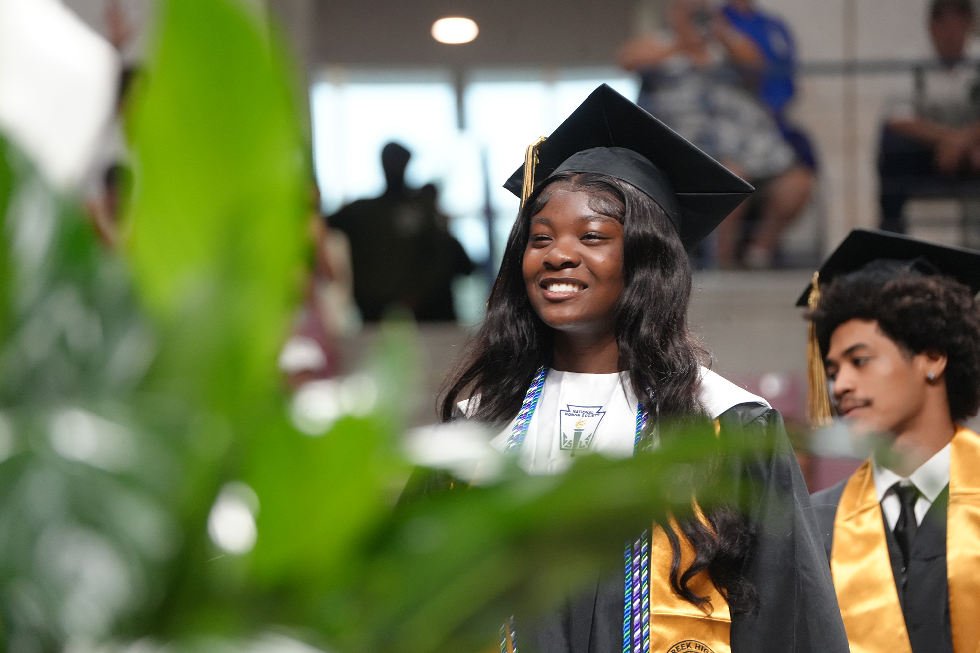 A smiling woman in a graduation gown and cap stands in front of a green plant. A man in a similar outfit stands behind her.