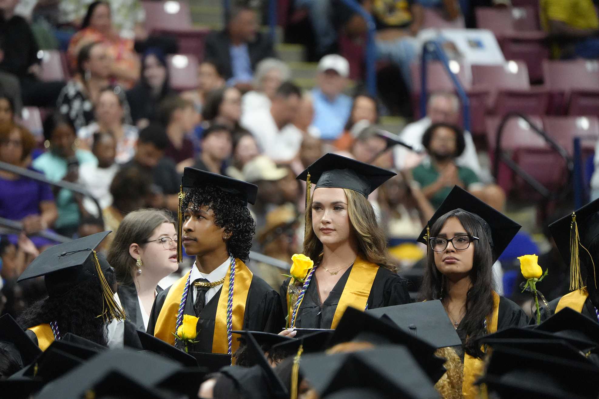 Graduates in caps and gowns stand in a row, facing the audience in a stadium. Some hold yellow flowers.