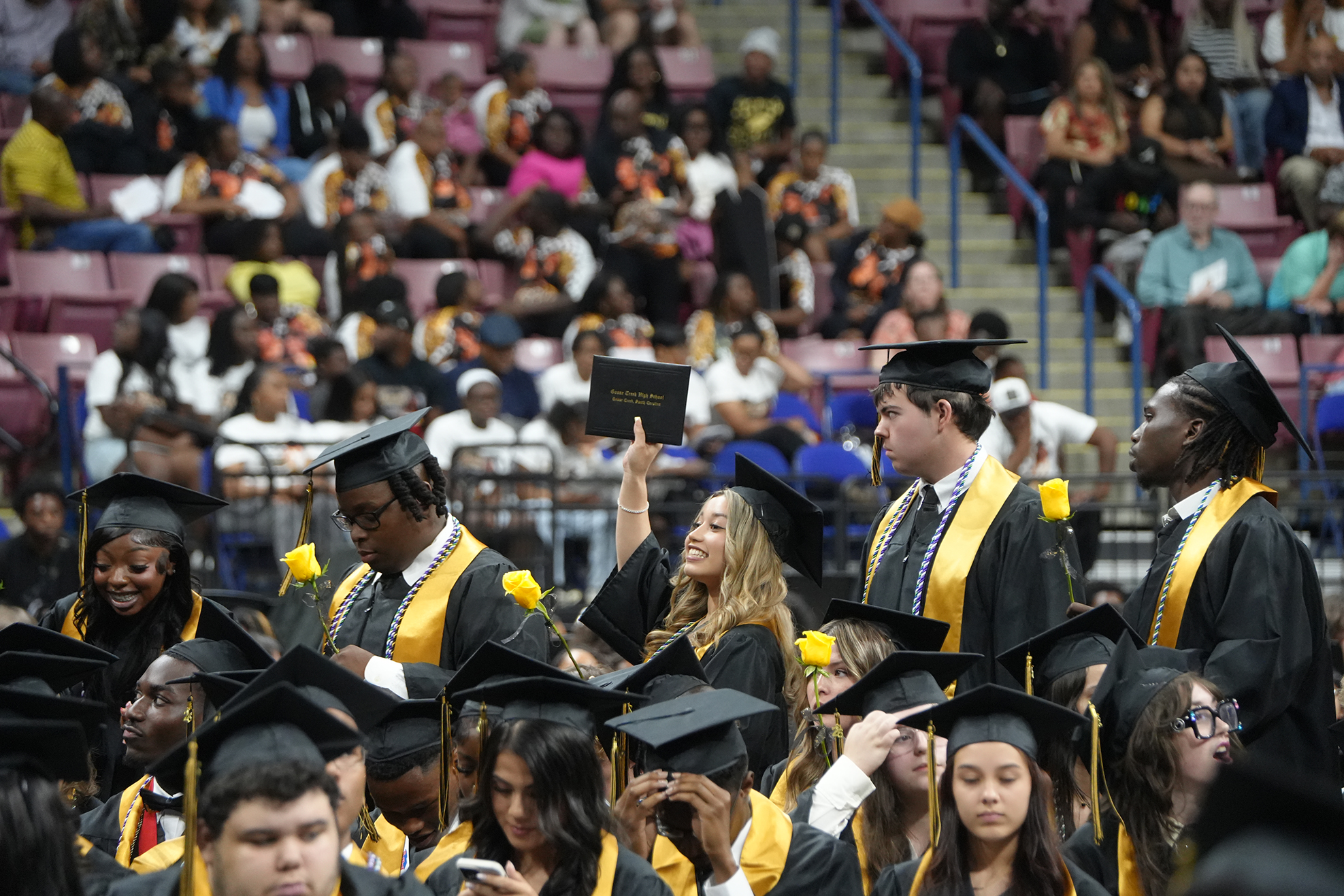 A crowd of people in graduation gowns, some holding up diplomas, stand in rows. Audience members sit in bleachers behind them.