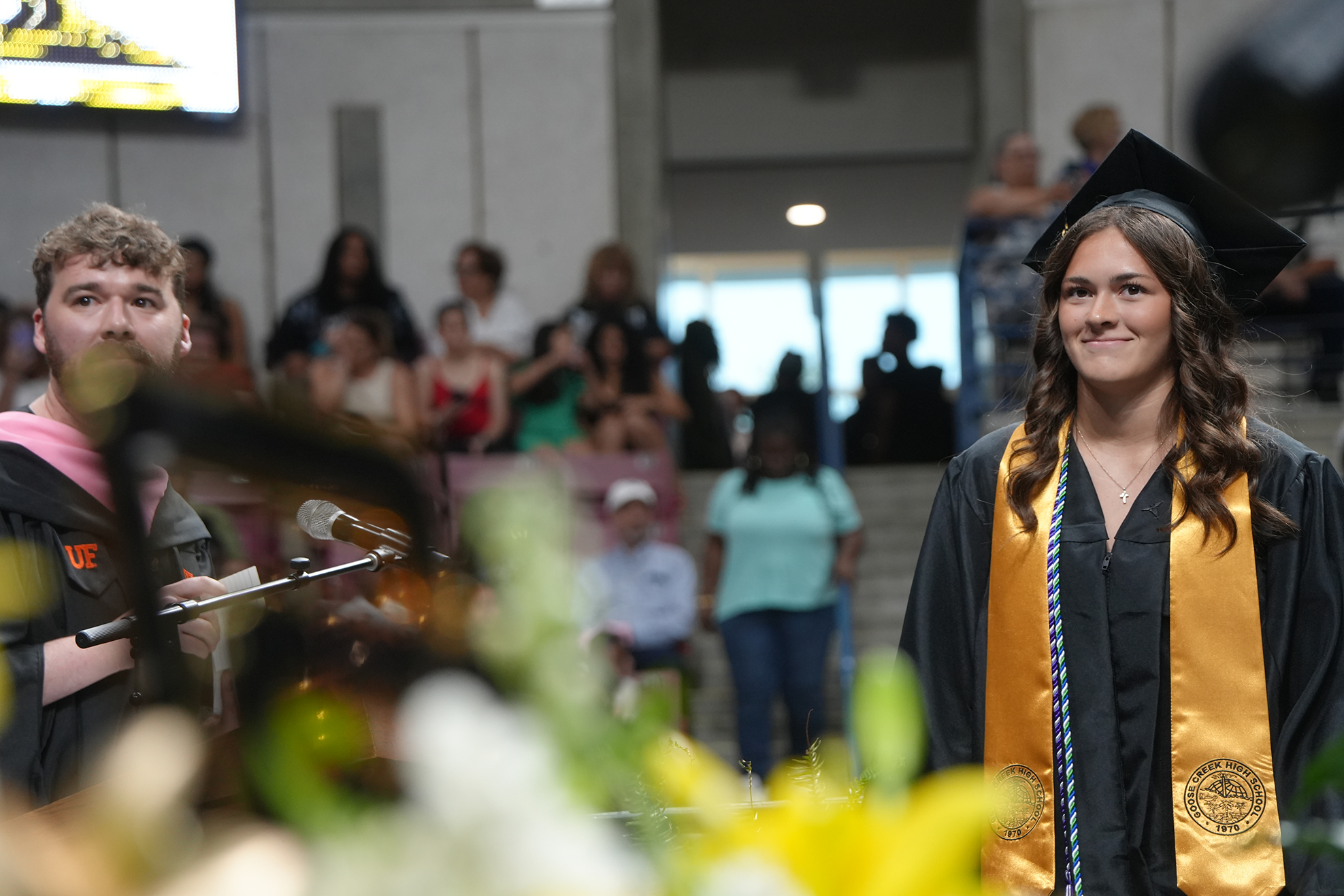 A woman in a graduation gown stands next to a man at a graduation ceremony, with people seated in the background.