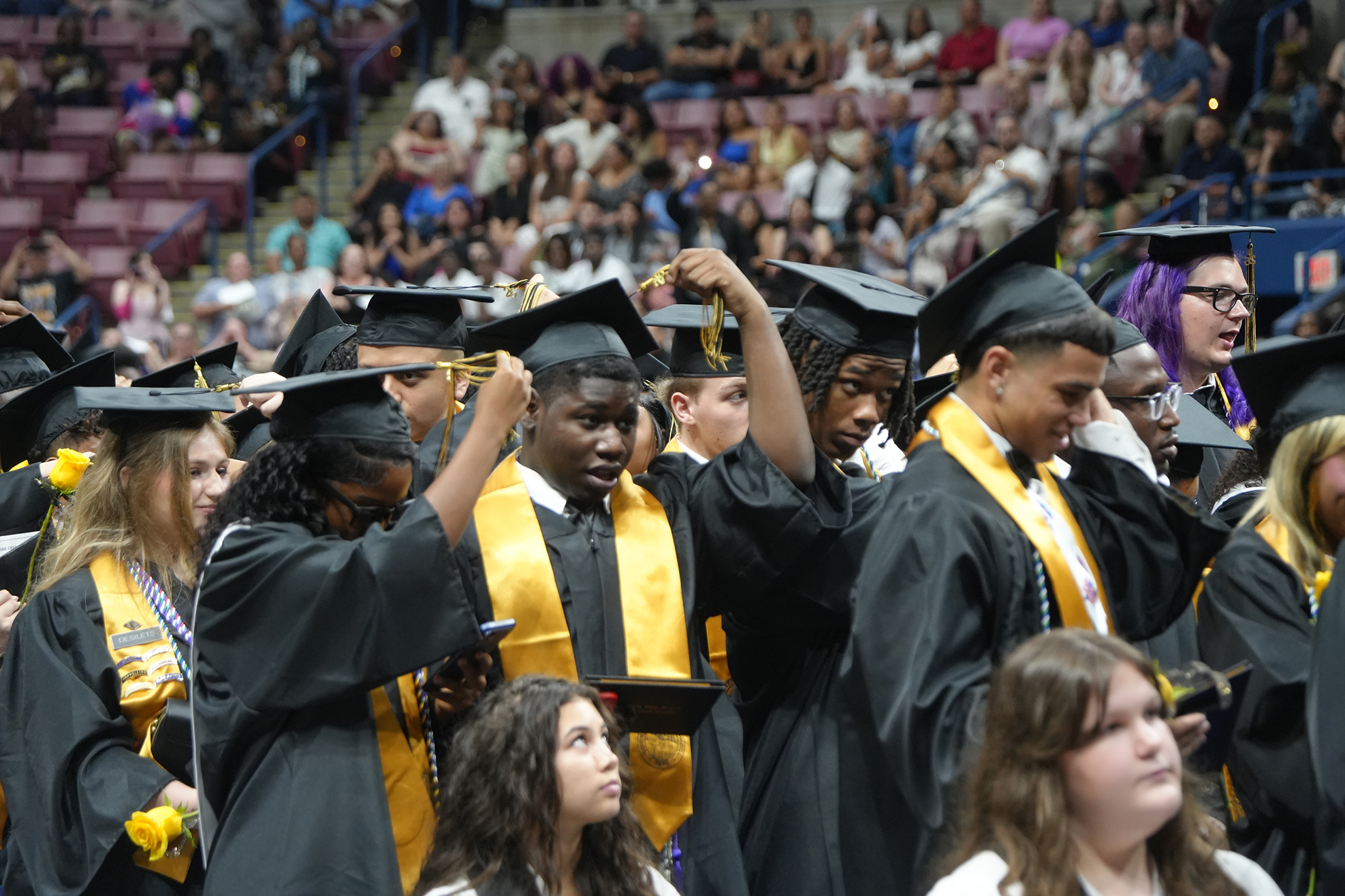 People in black and yellow graduation gowns and caps standing in a stadium during a ceremony.