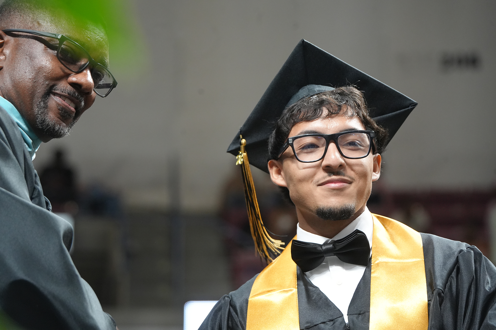 A man in a cap and gown shakes hands with another person. Both wear glasses. Behind them, a blurred background with seating.