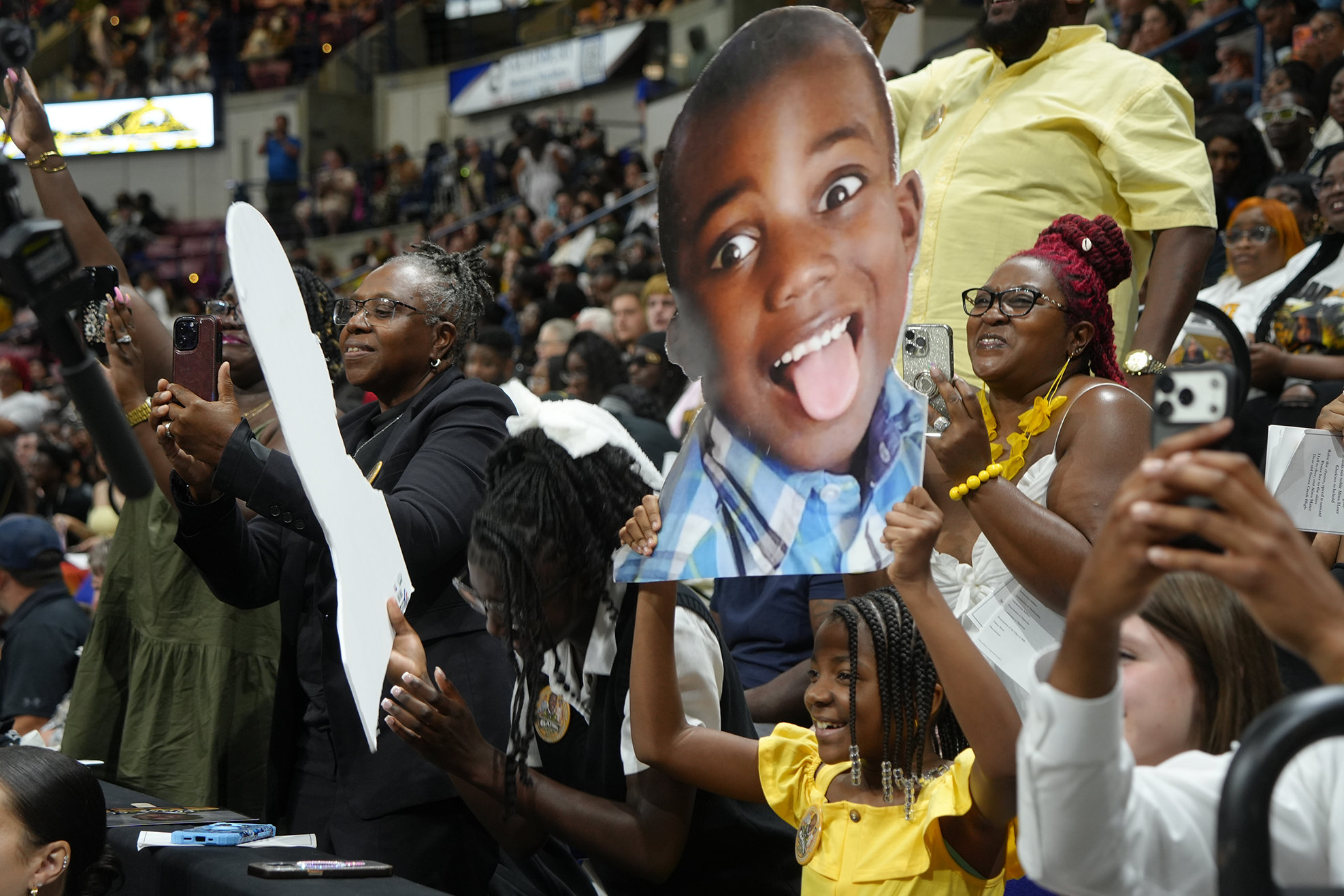 Group of people hold up signs and a photo cutout of a smiling child at a stadium.