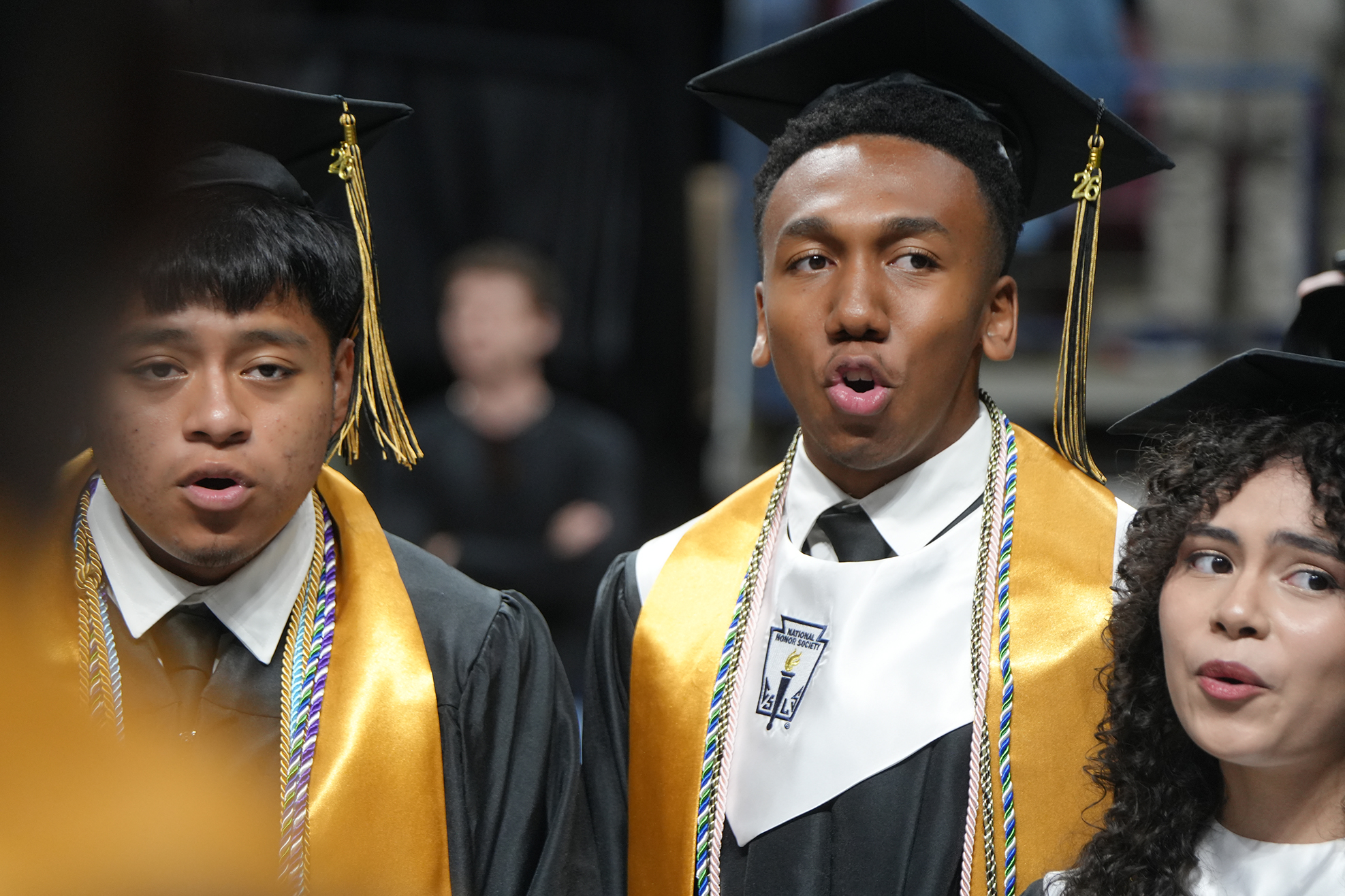Three individuals in graduation attire. One man is speaking, another man is listening, and a woman is looking up.