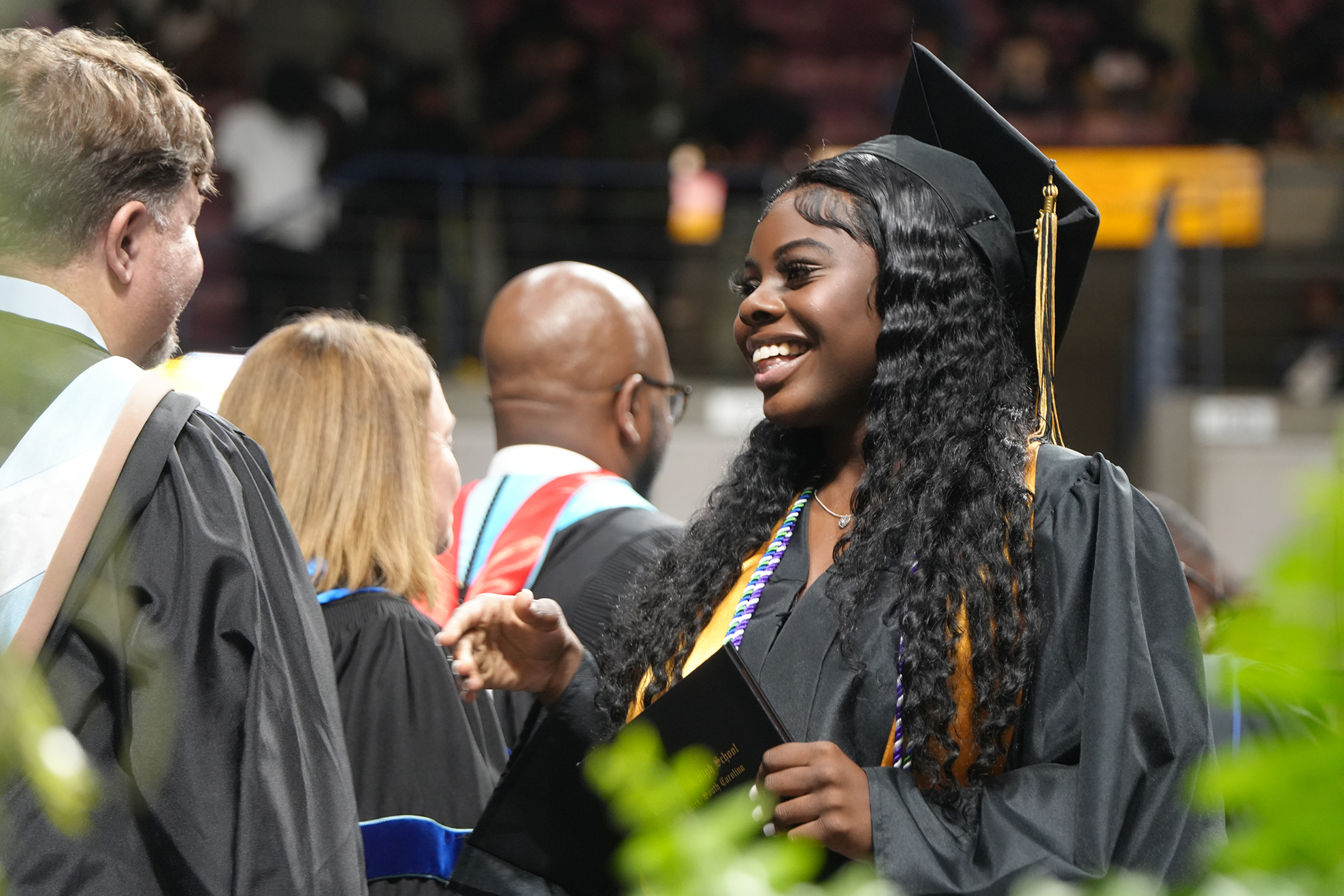 A smiling woman in a black graduation gown stands among other graduates at an outdoor graduation ceremony.
