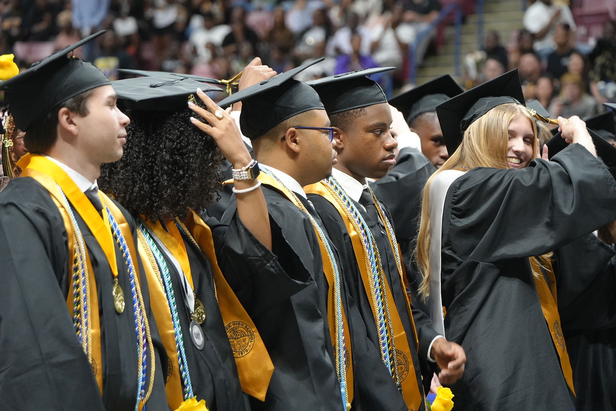 Graduates in black gowns and caps, some with tassels, stand in a row. A blurred audience in the background.