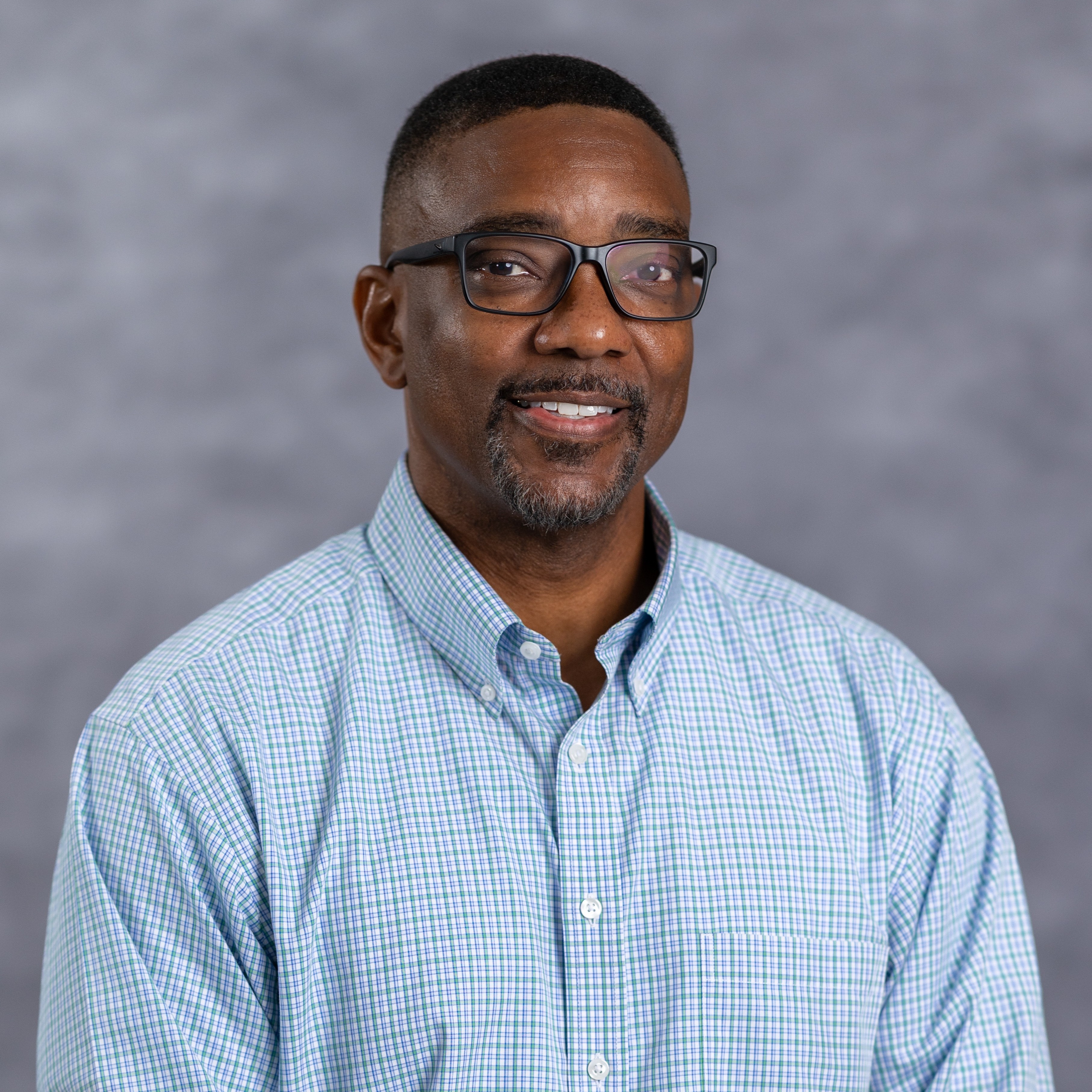 A man in a light blue checked shirt and glasses stands against a gray background, smiling.