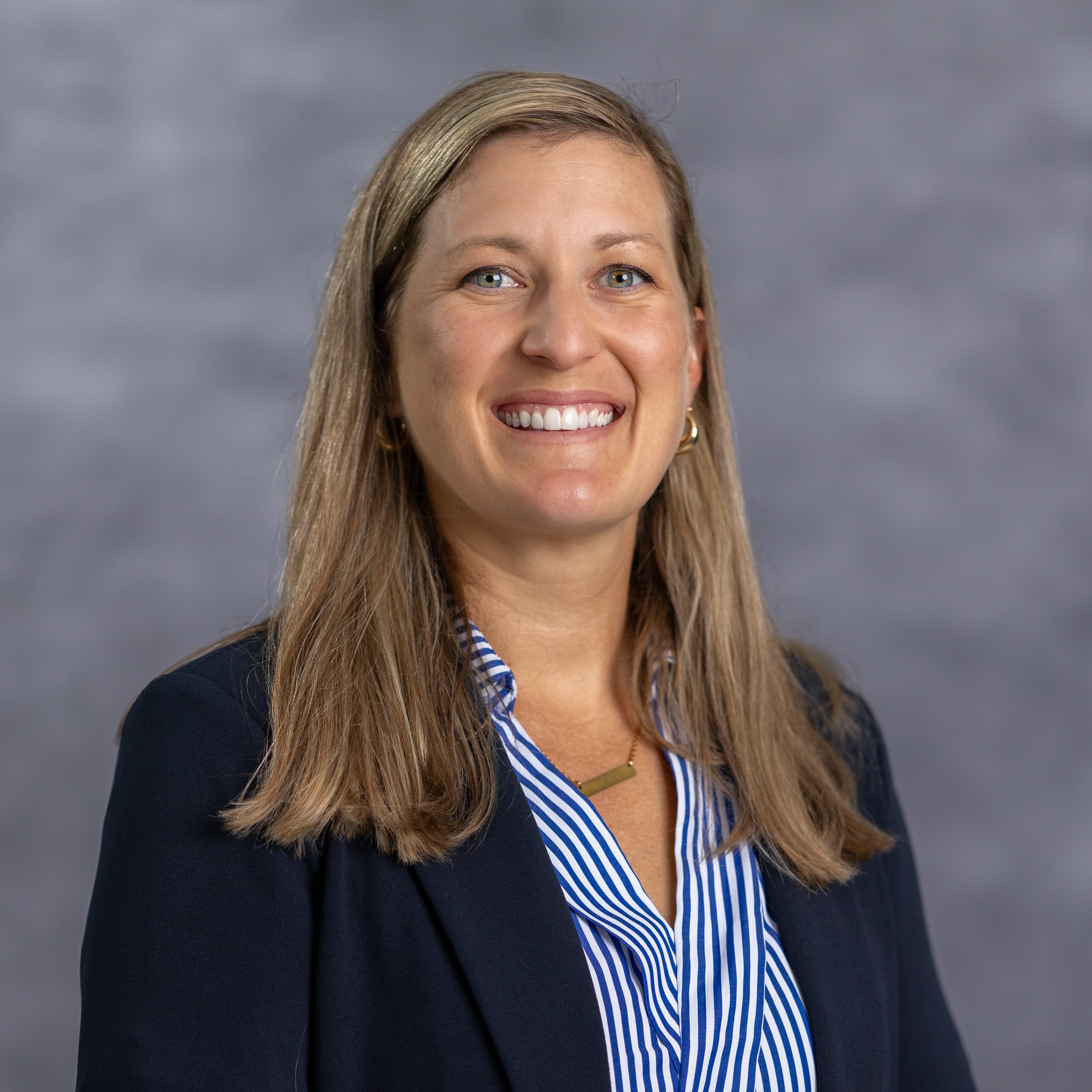 A woman with blonde hair smiling and wearing a navy blazer over a blue and white striped shirt.