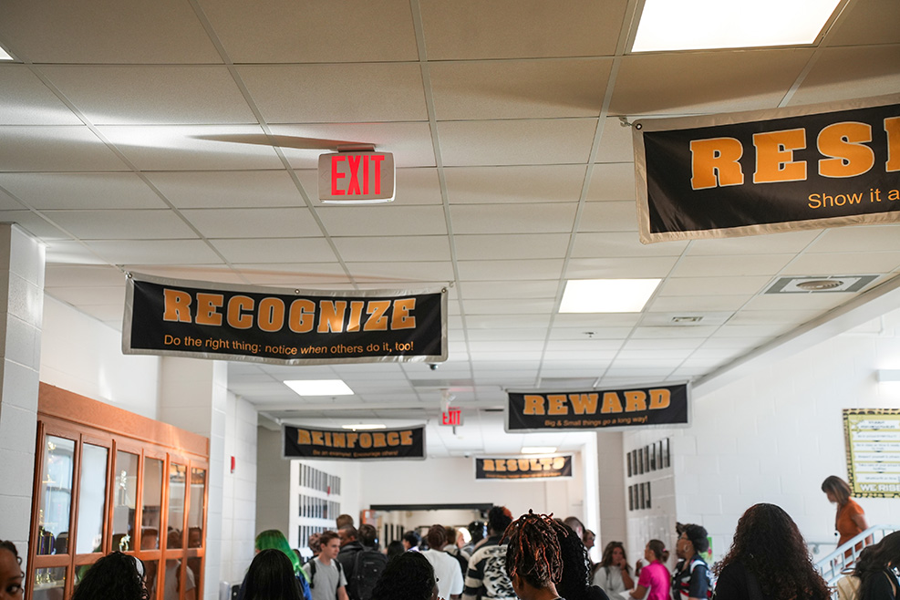Hallway with banners: "RES", "RECOGNIZE", "REWARD". People standing and moving. White walls, ceiling lights.