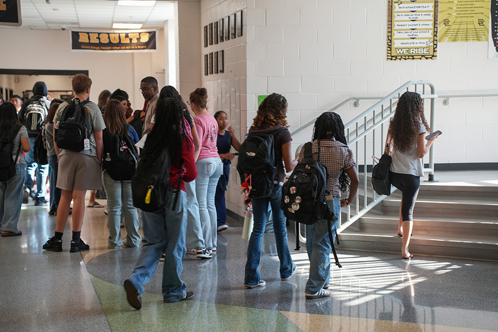 Students with backpacks in a school hallway, some standing in line and others descending a staircase.