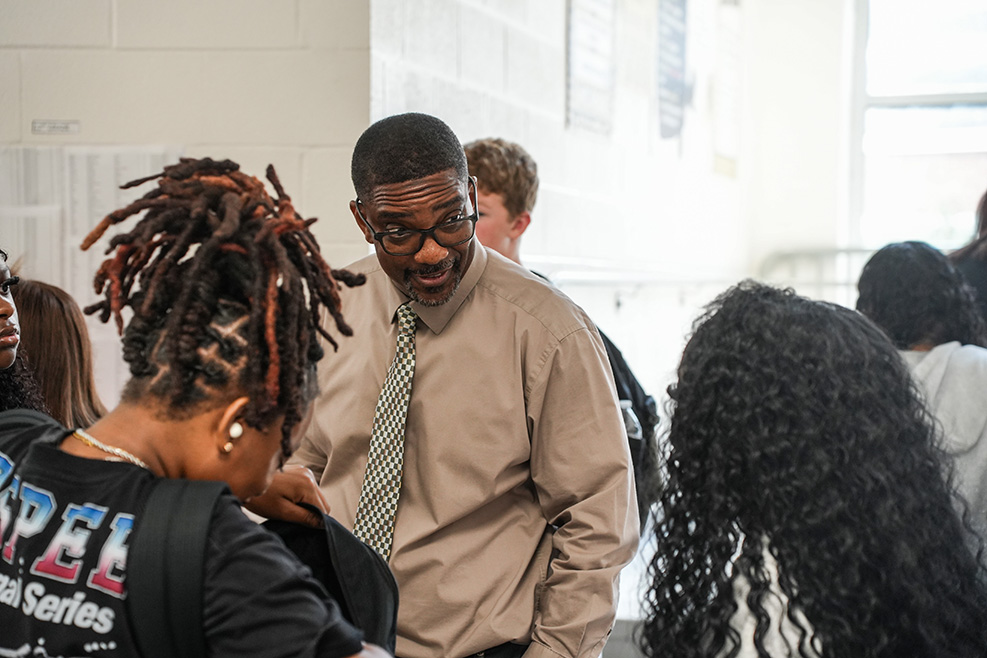A person with a long braid speaks with a man in a tan shirt, tie, and glasses.