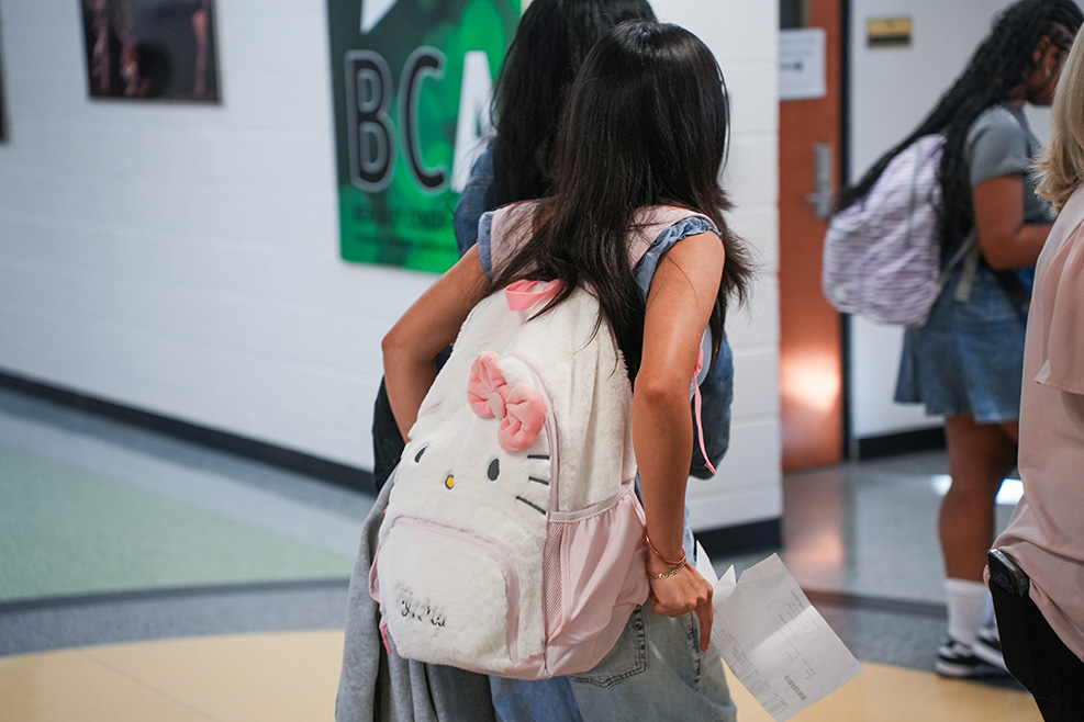 Students walk through a hallway. A girl has a Hello Kitty backpack. A girl with a blue dress walks beside her.