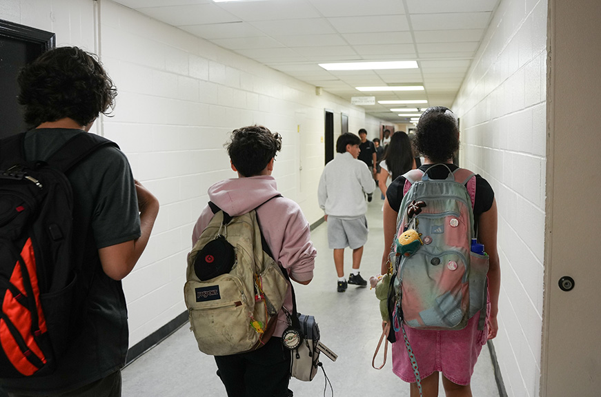 People walking in a school hallway. Some are carrying backpacks, one wears a pink hoodie.