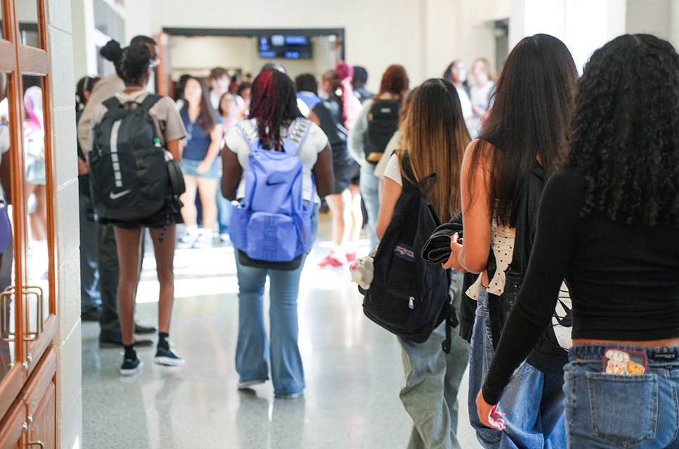 A hallway with people wearing backpacks. Some wear jeans, others skirts. The floor reflects light.