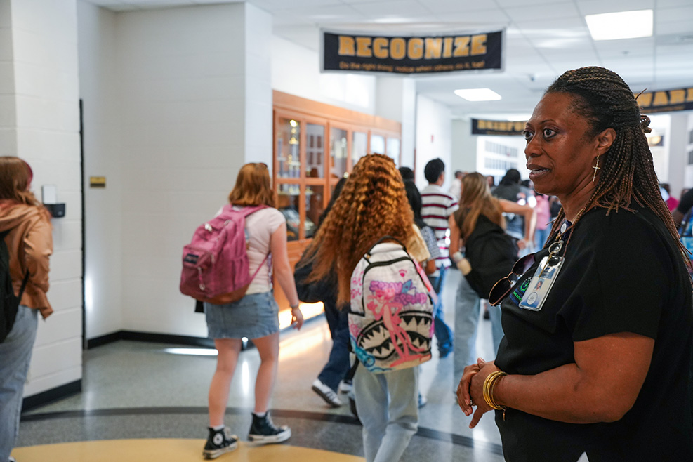 A hallway with a banner reading "RECOGNIZE" above. A woman in black speaks to another woman with a backpack.
