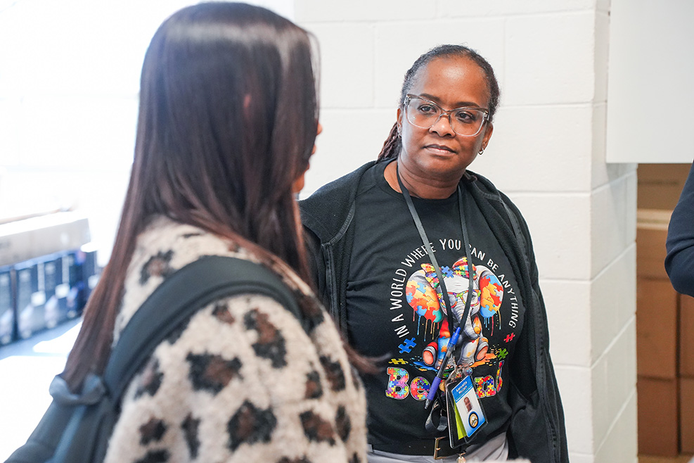 Two women talk, one with a leopard-print jacket and backpack, the other wearing glasses and a black shirt with a colorful design.