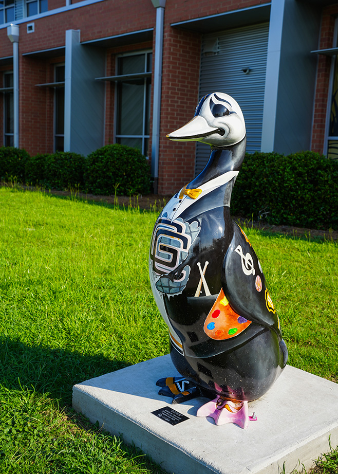 A large duck statue on display outdoors. It has colorful designs and is on a concrete block. A brick building and shrubs are in the background.