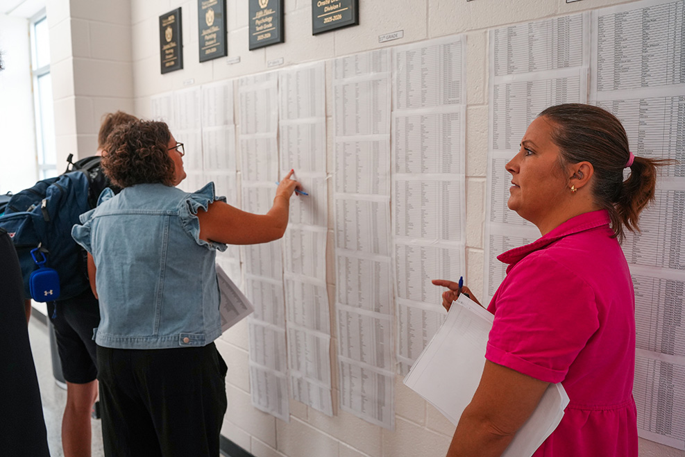 Two women read a bulletin board with many typed pages pinned to it. One holds a marker.