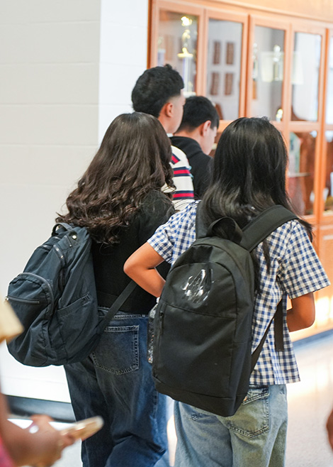 Students with backpacks stand in a hallway. A display case with glass doors is behind them.