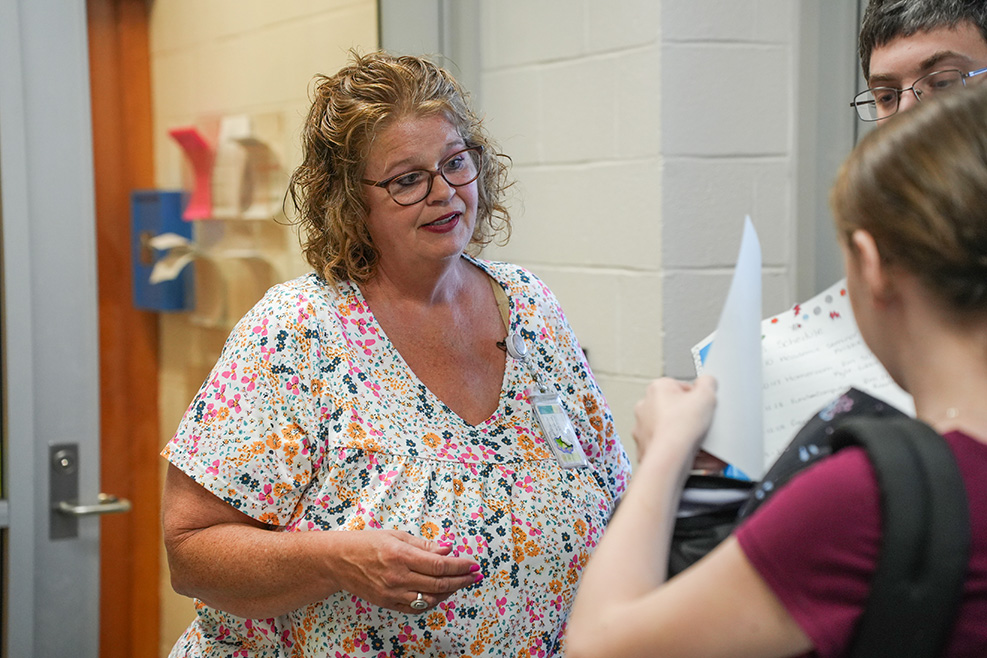 A woman with glasses and a floral top speaks to a student holding papers and a backpack.