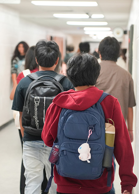 Students in a school hallway, one with a blue backpack and a red hoodie, another with a black backpack.