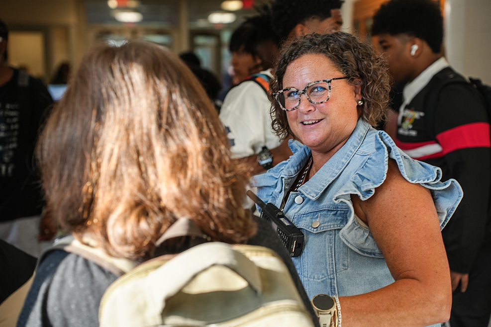 An adult woman with curly hair and glasses speaks to a person with a backpack in a hallway.