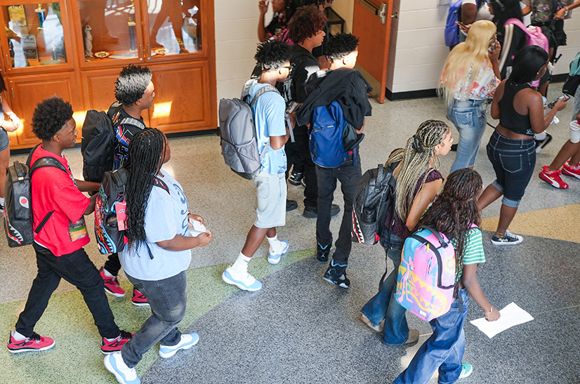 A hallway with students walking. Some carry backpacks. One holds a piece of paper.