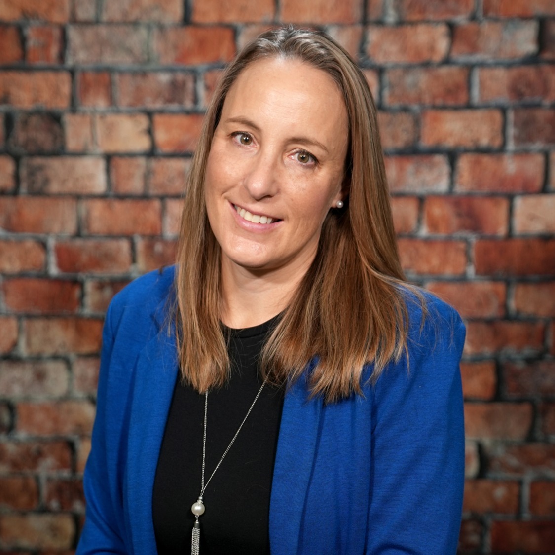 Woman wearing a blue blazer and black shirt smiles, with a necklace, standing before a brick wall.