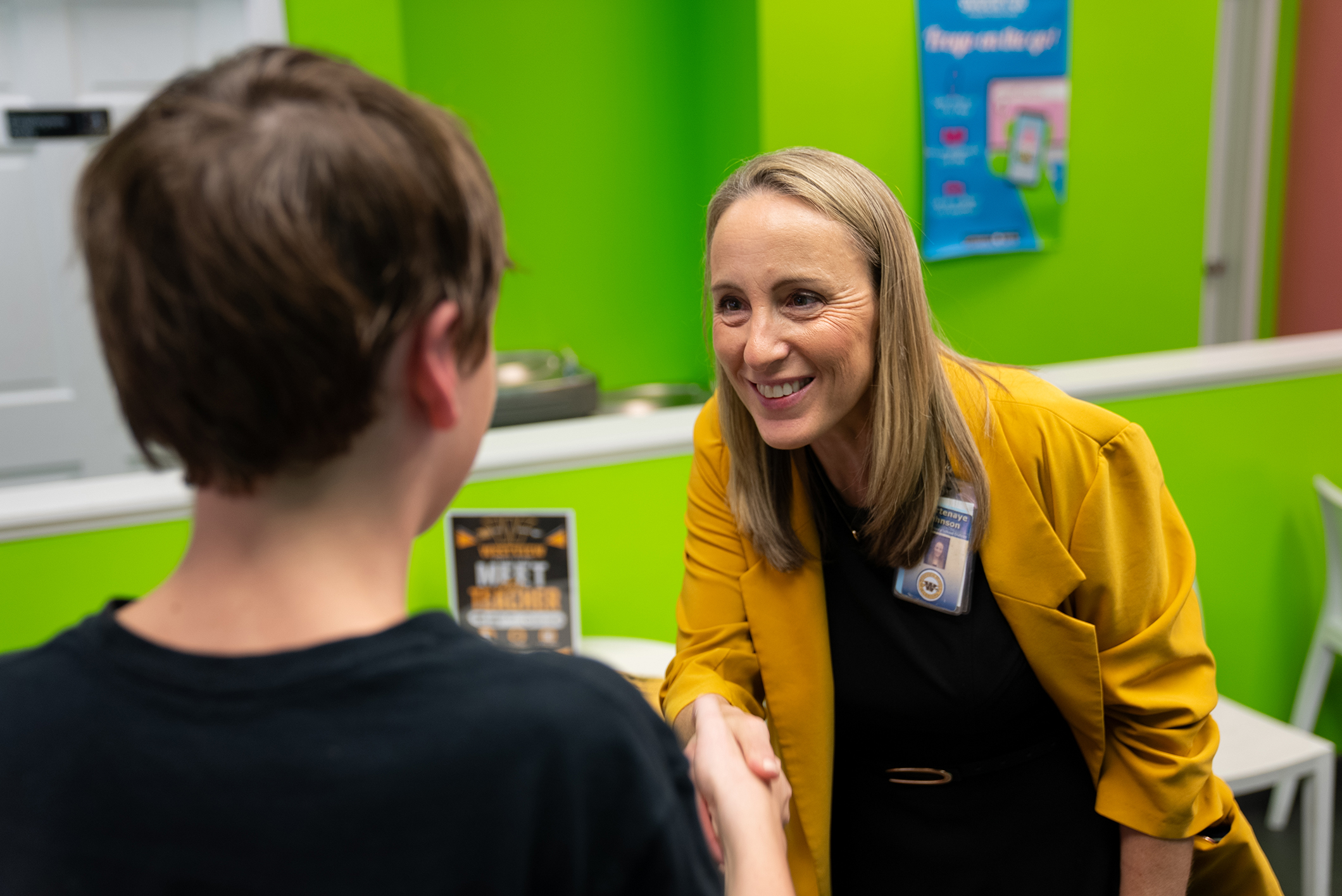 A woman with blonde hair wearing a yellow jacket shakes hands with a boy in a black shirt.