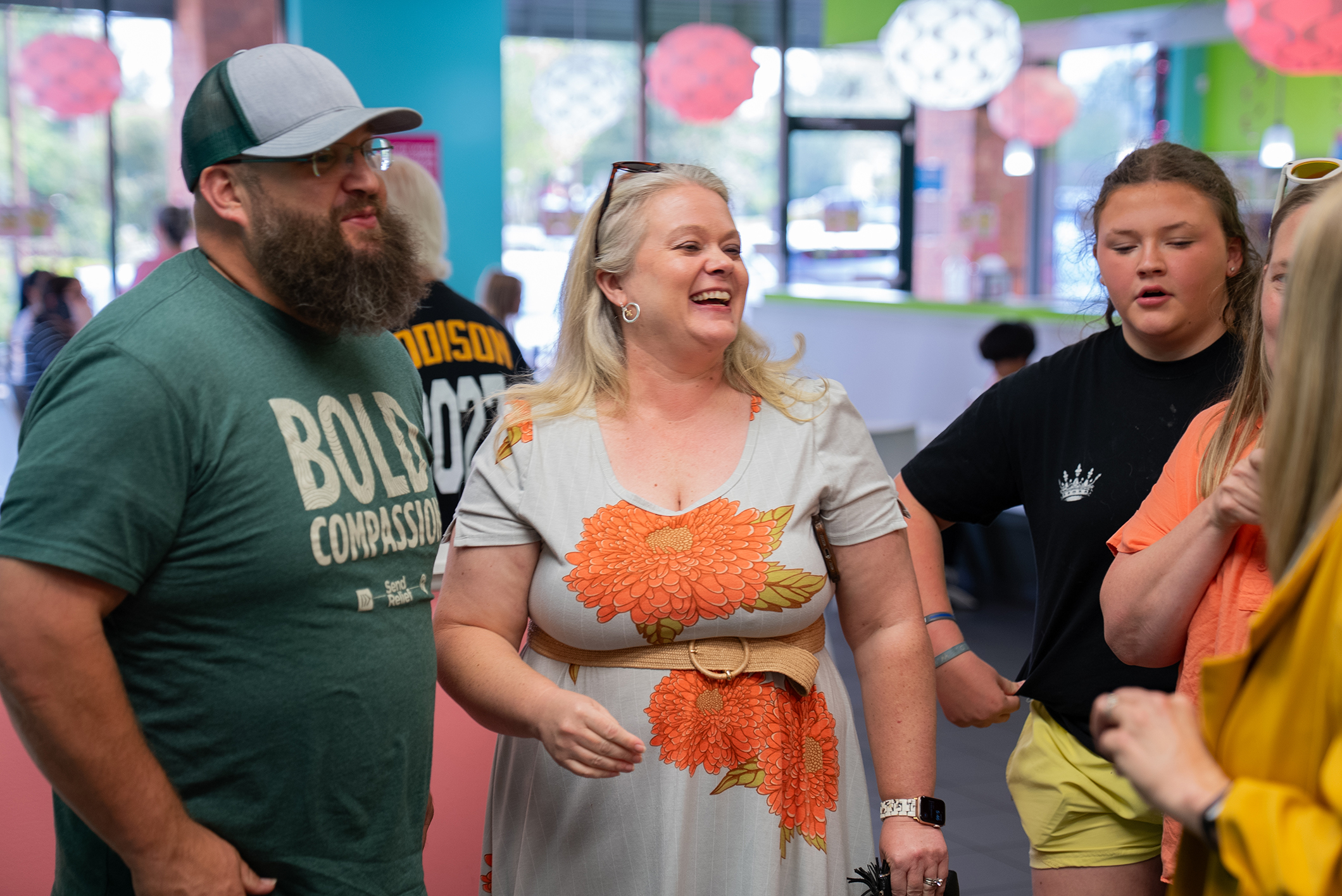 People interact in a room with bright colors. A woman in a floral dress laughs. A man in a green shirt stands next to her.