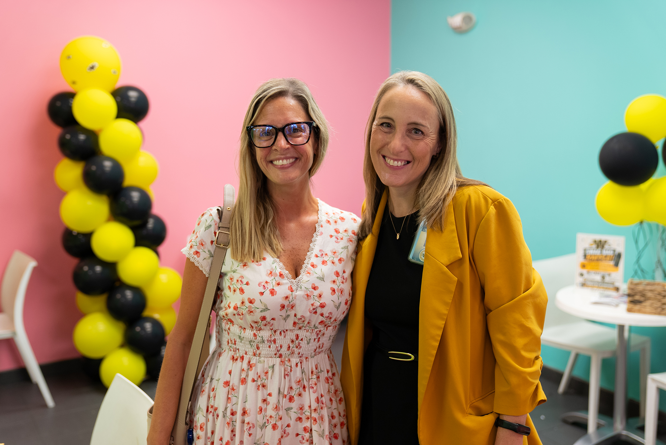 Two women stand next to each other in a room with colorful walls and balloons. One wears a floral dress, and the other wears a yellow coat.