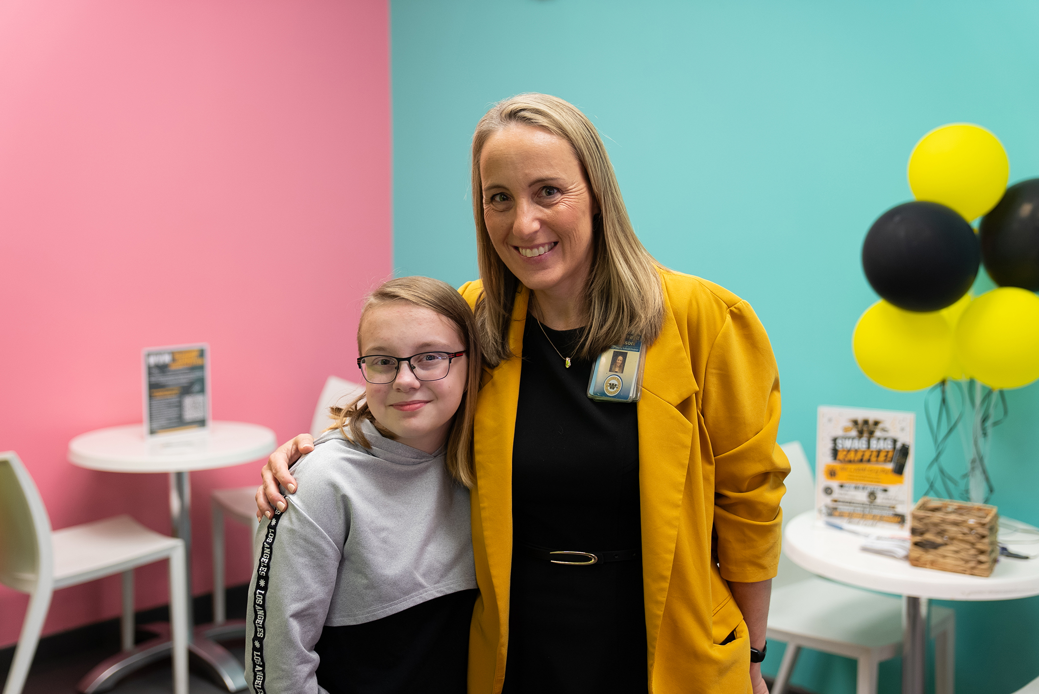 Two women pose for a photo in a room with pastel-colored walls, balloons, and tables with chairs.