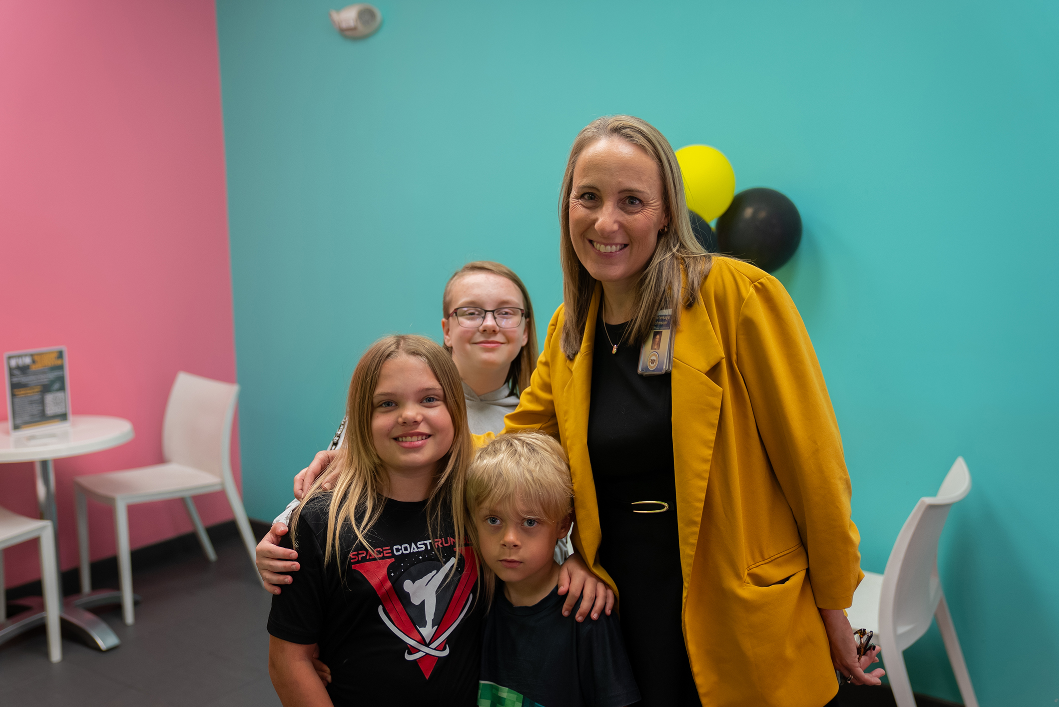 Four individuals, two women and two children, smiling and posing in a colorful room with balloons and chairs.