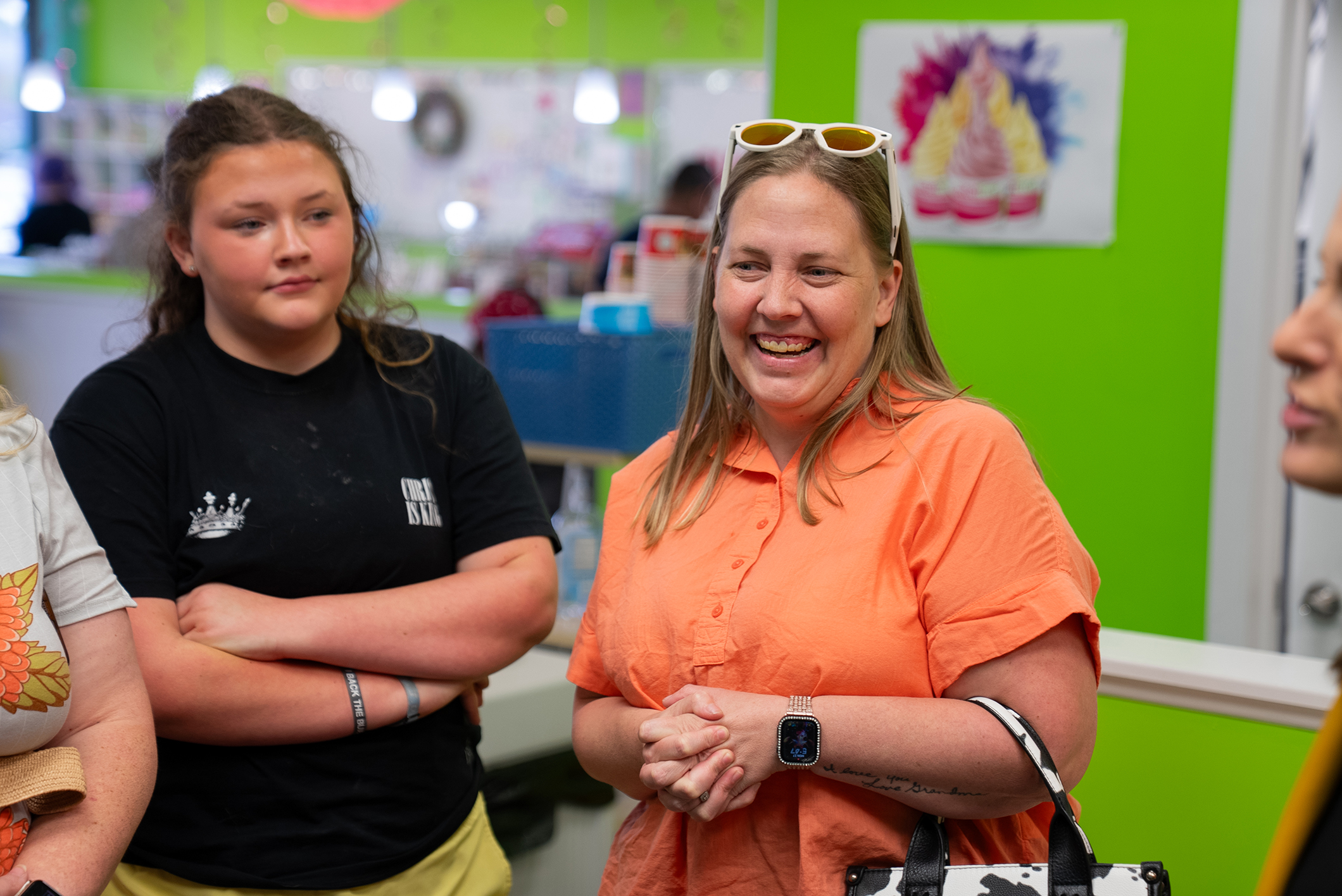 Three women stand in a brightly lit room; one wears an orange shirt, another a black shirt, and the third has folded arms. A green wall with a decorative item is behind them.