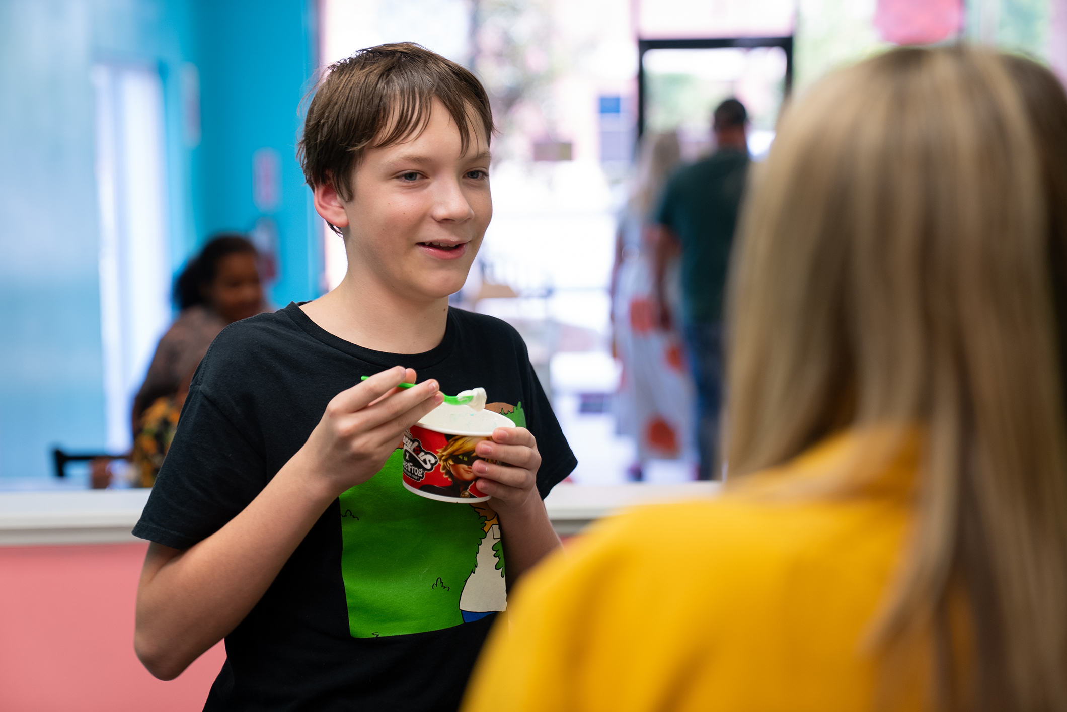 A boy holds a bowl of food while speaking to a woman; blurry people in a colorful room in the background.