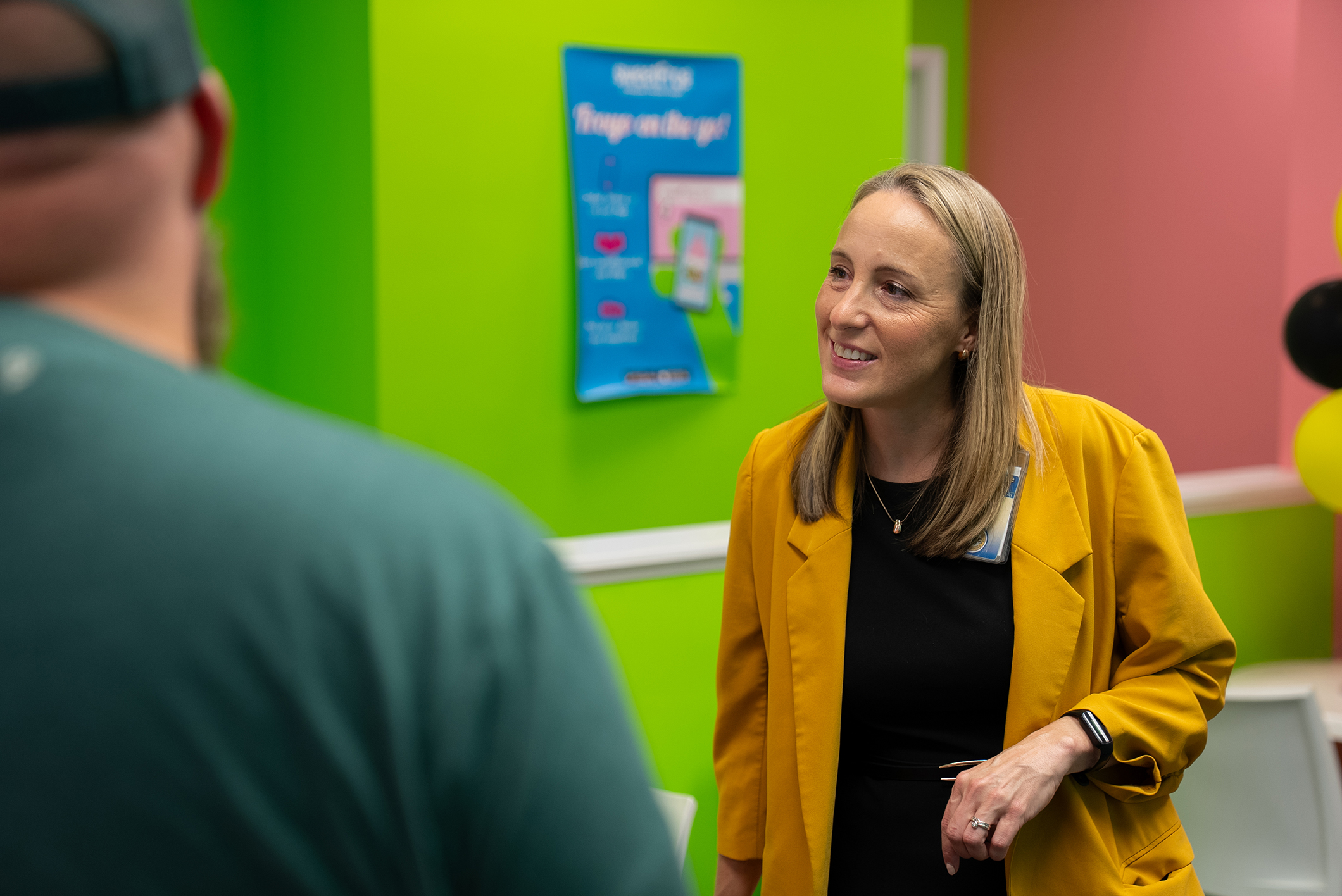 Woman in a yellow jacket speaking with a man in a green shirt. Green and pink walls with balloons.