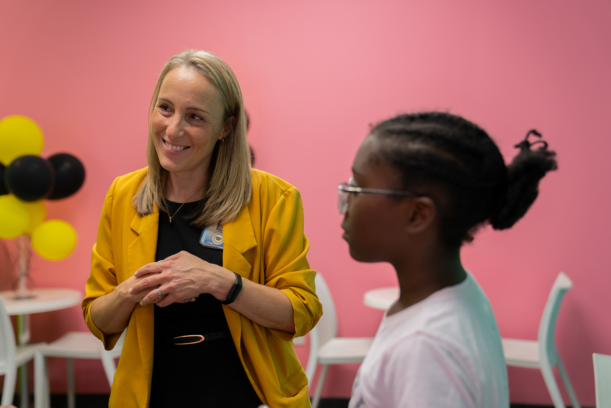 A woman in a yellow jacket talks with a young girl in a white shirt in a brightly colored room.