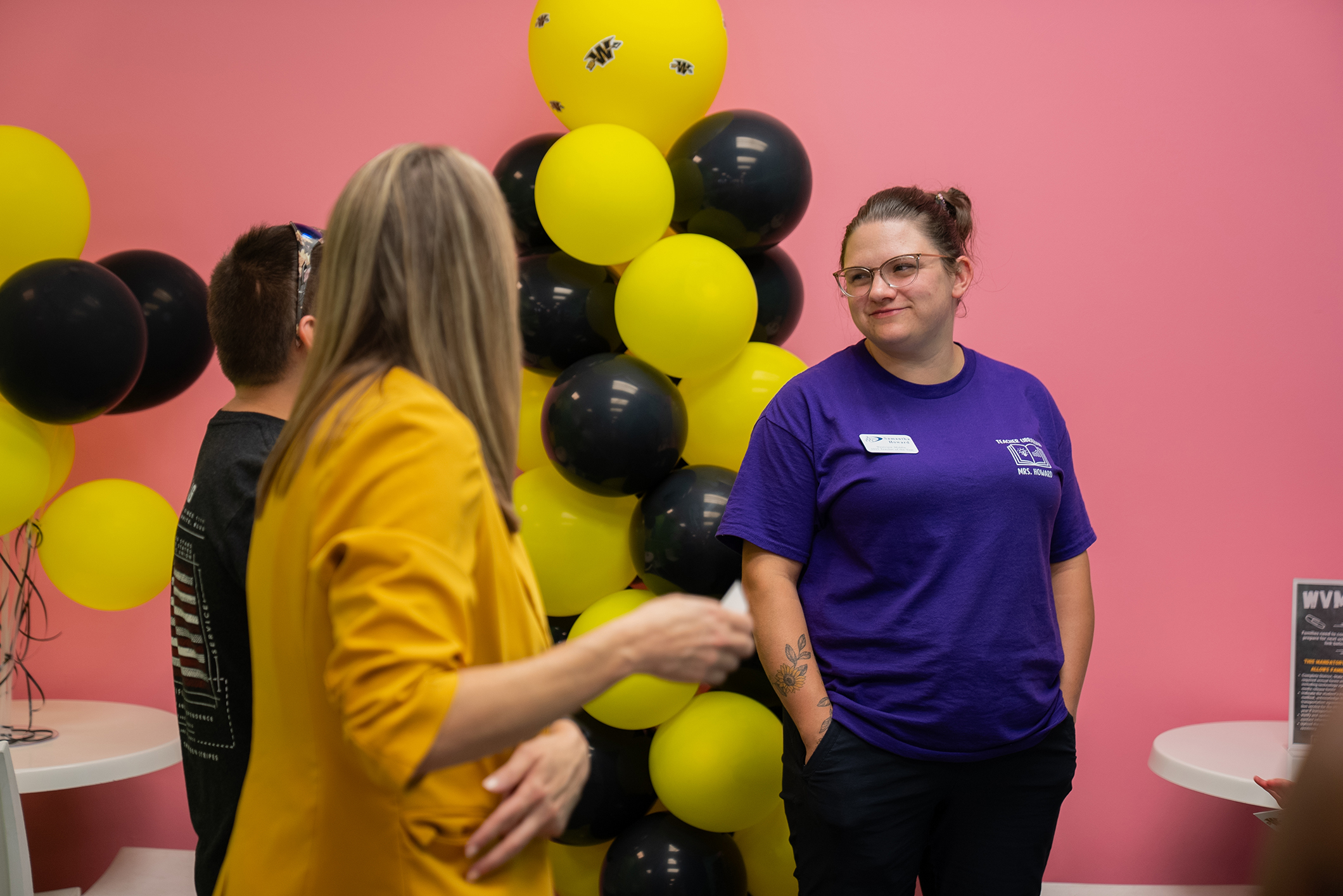 Two women stand next to a wall with yellow and black balloons. One wears a yellow blazer, the other a purple shirt.