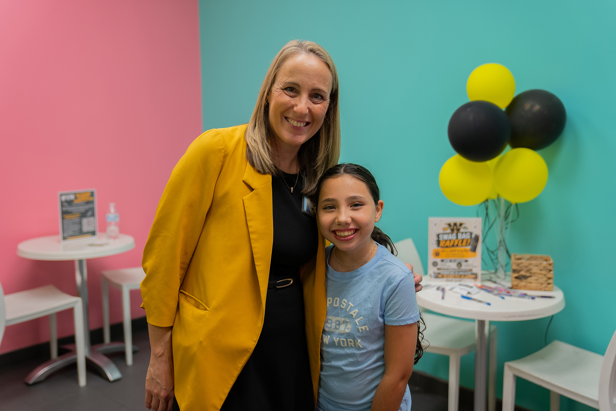 A woman and girl stand in a room with colorful walls. The woman wears a yellow jacket; the girl wears a blue shirt.