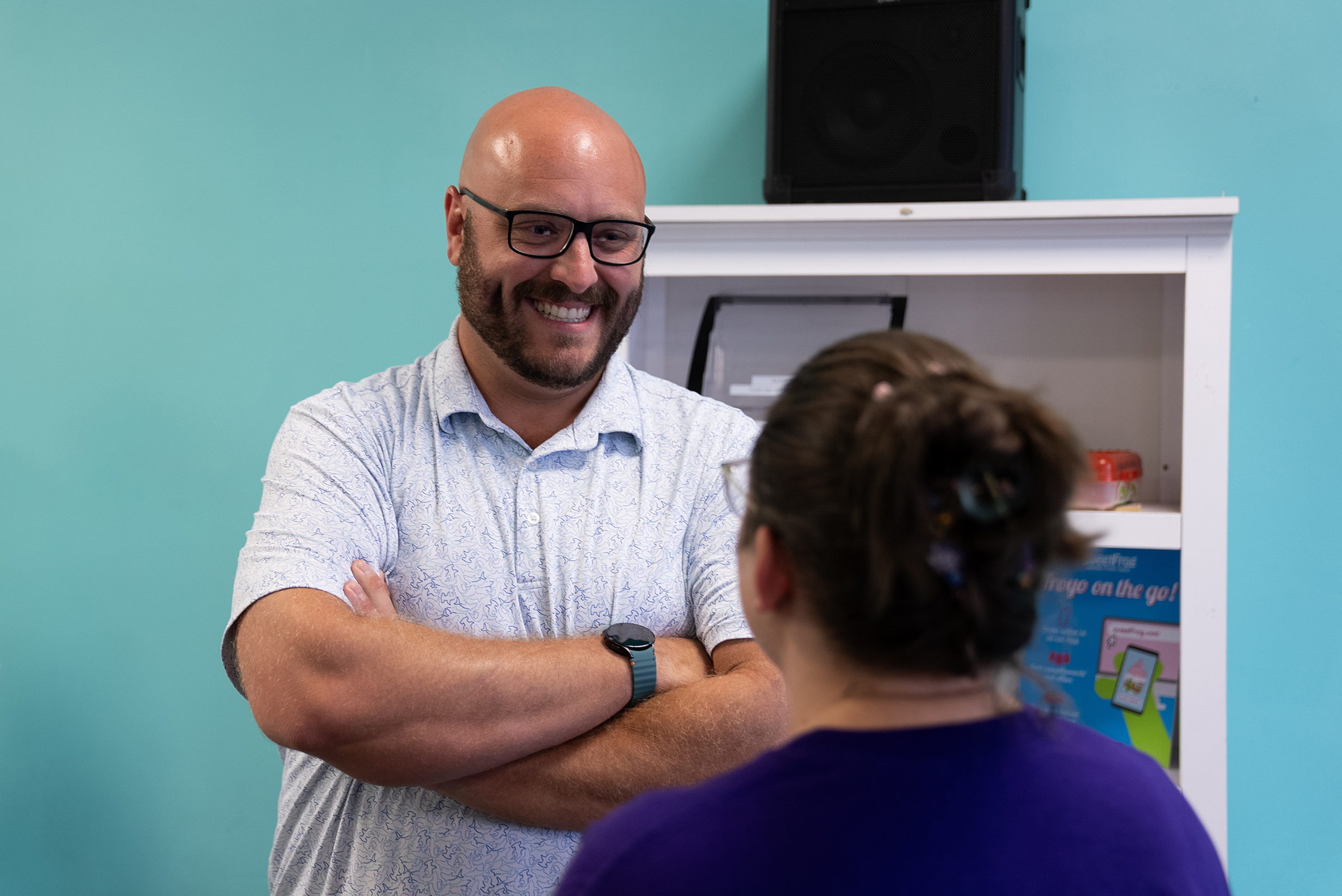 Man with glasses and watch talks to woman with ponytail. Background has a white shelf, blue wall, and speaker.