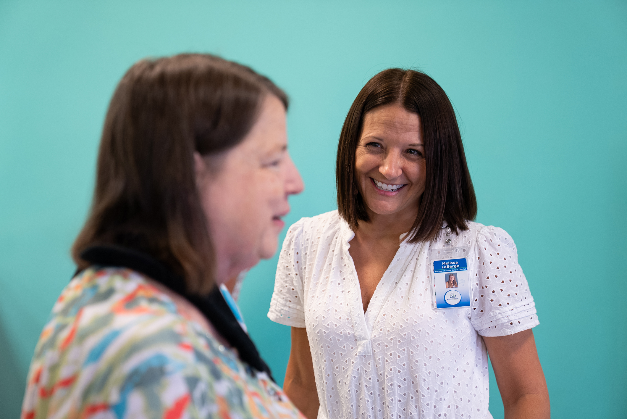 Two women converse in a room with a teal wall; one wears a white blouse with a name tag.