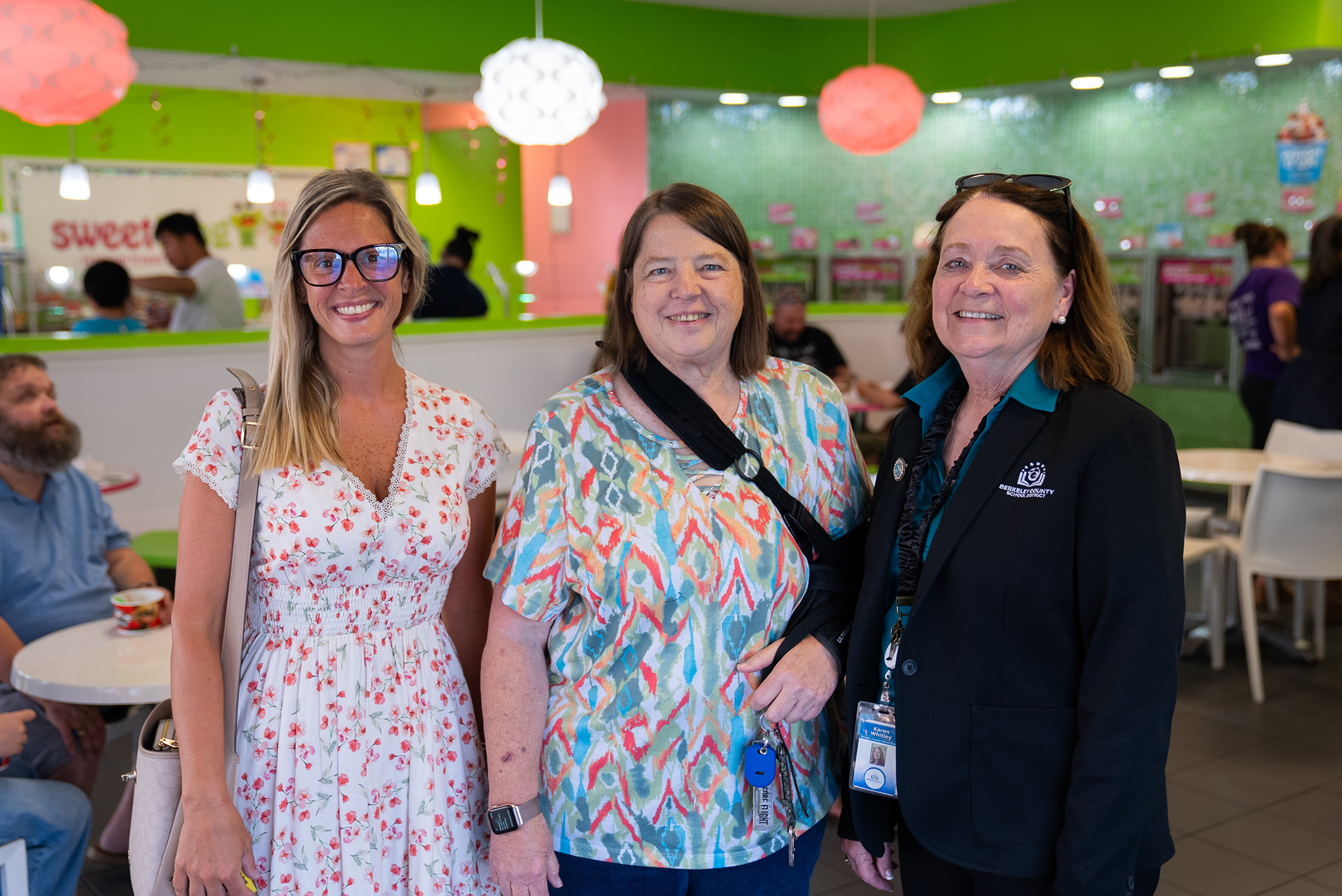 Three women, one in floral dress, one with a camera, and one in black jacket, smiling. Behind them, a blurry view of a dining area with green walls.