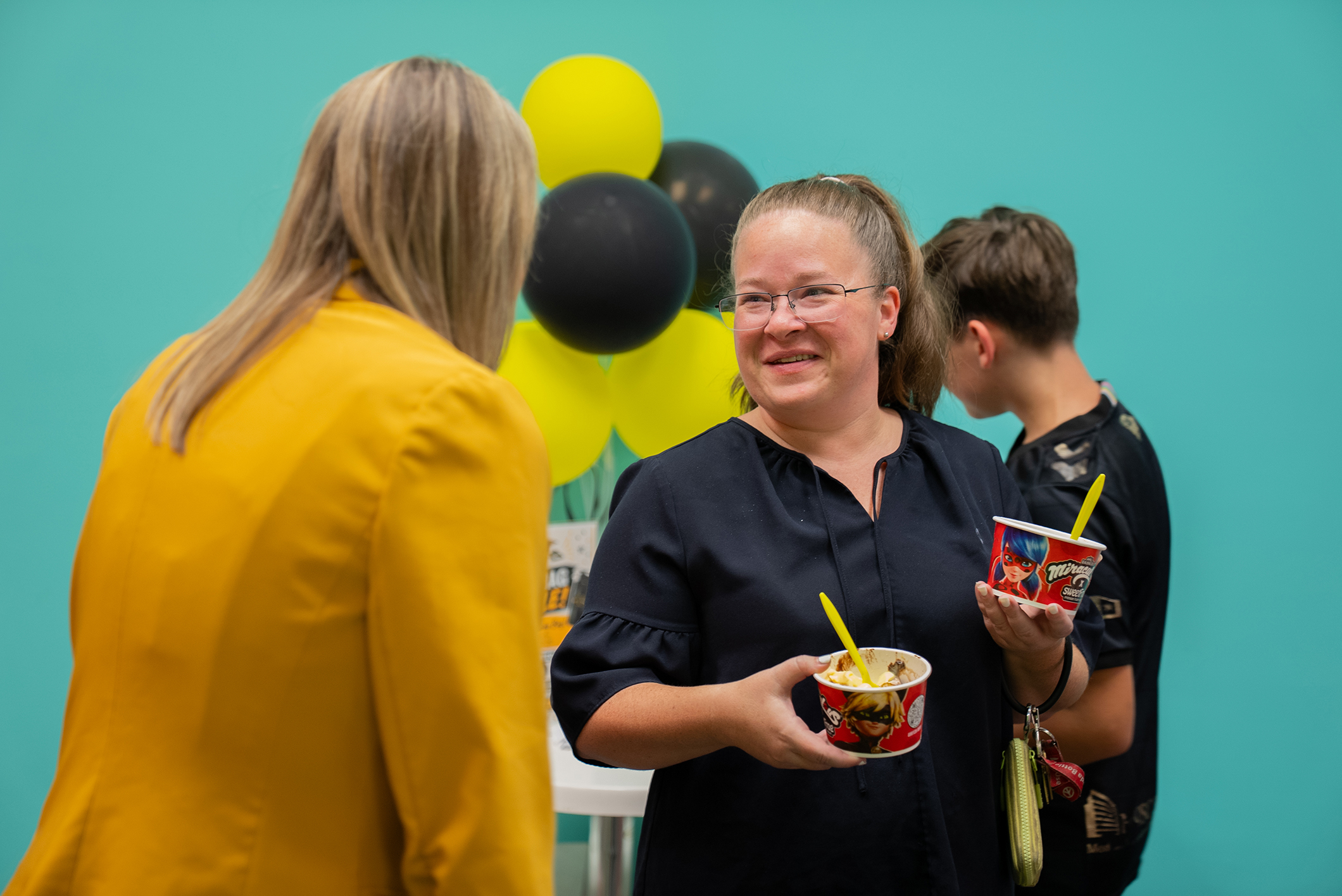 Three people gather with food cups in hand; one woman wears eyeglasses and a black shirt. Behind, a teal wall has balloons.