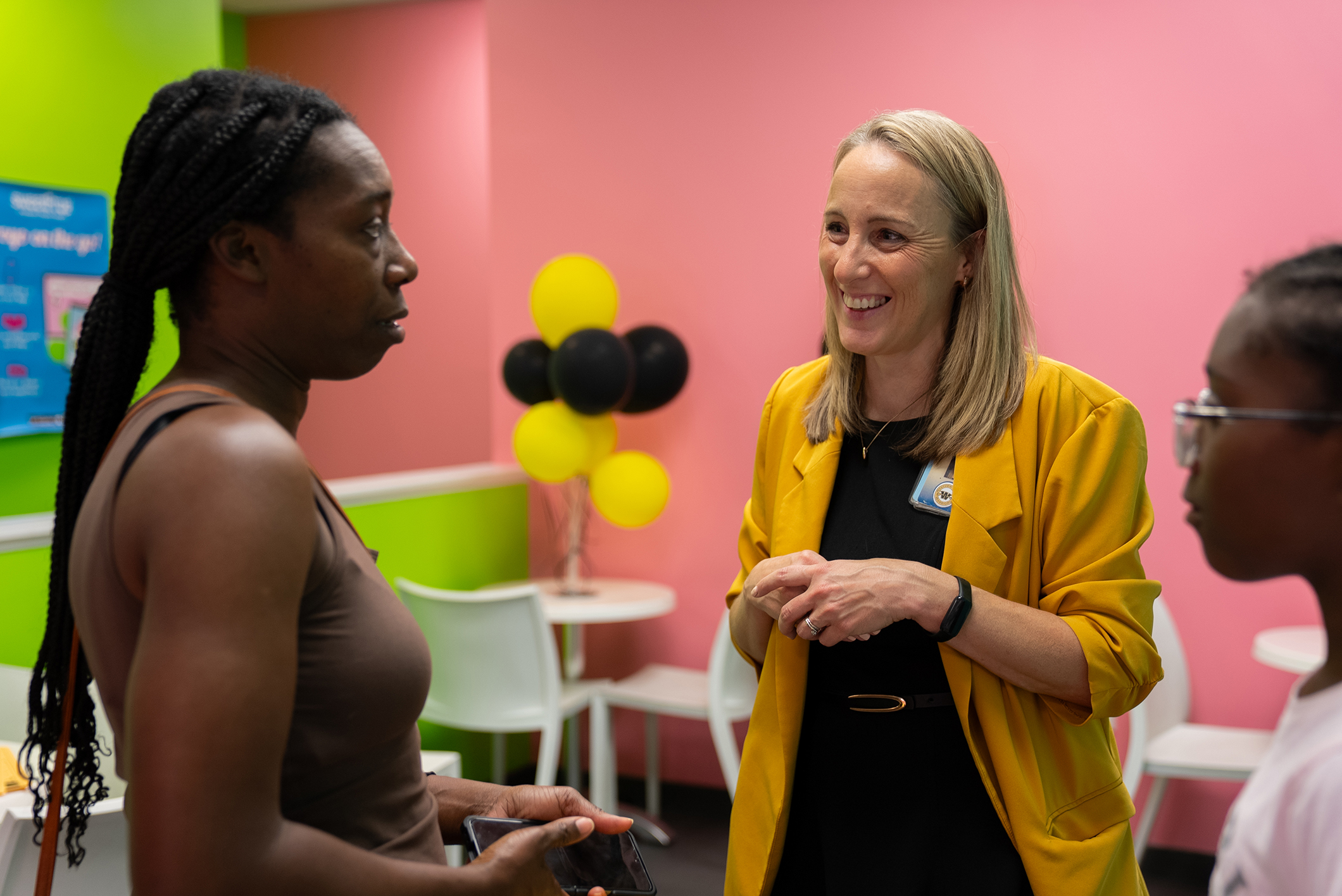 Three people stand in a room with pink and green walls. A woman in yellow speaks to another woman with braided hair. A child wearing glasses listens.