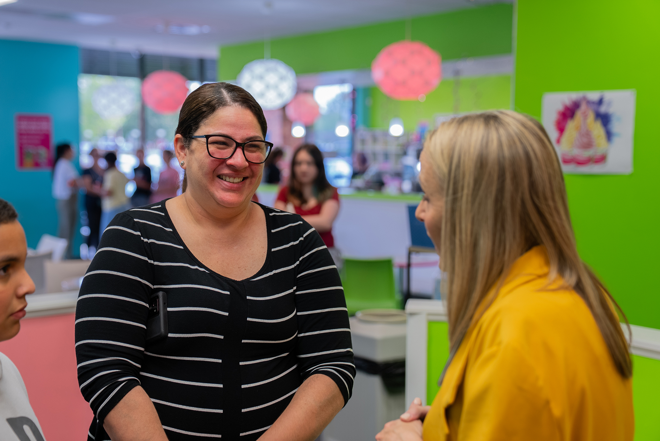Three women converse in a brightly colored room. The woman in the center wears glasses and a striped shirt.
