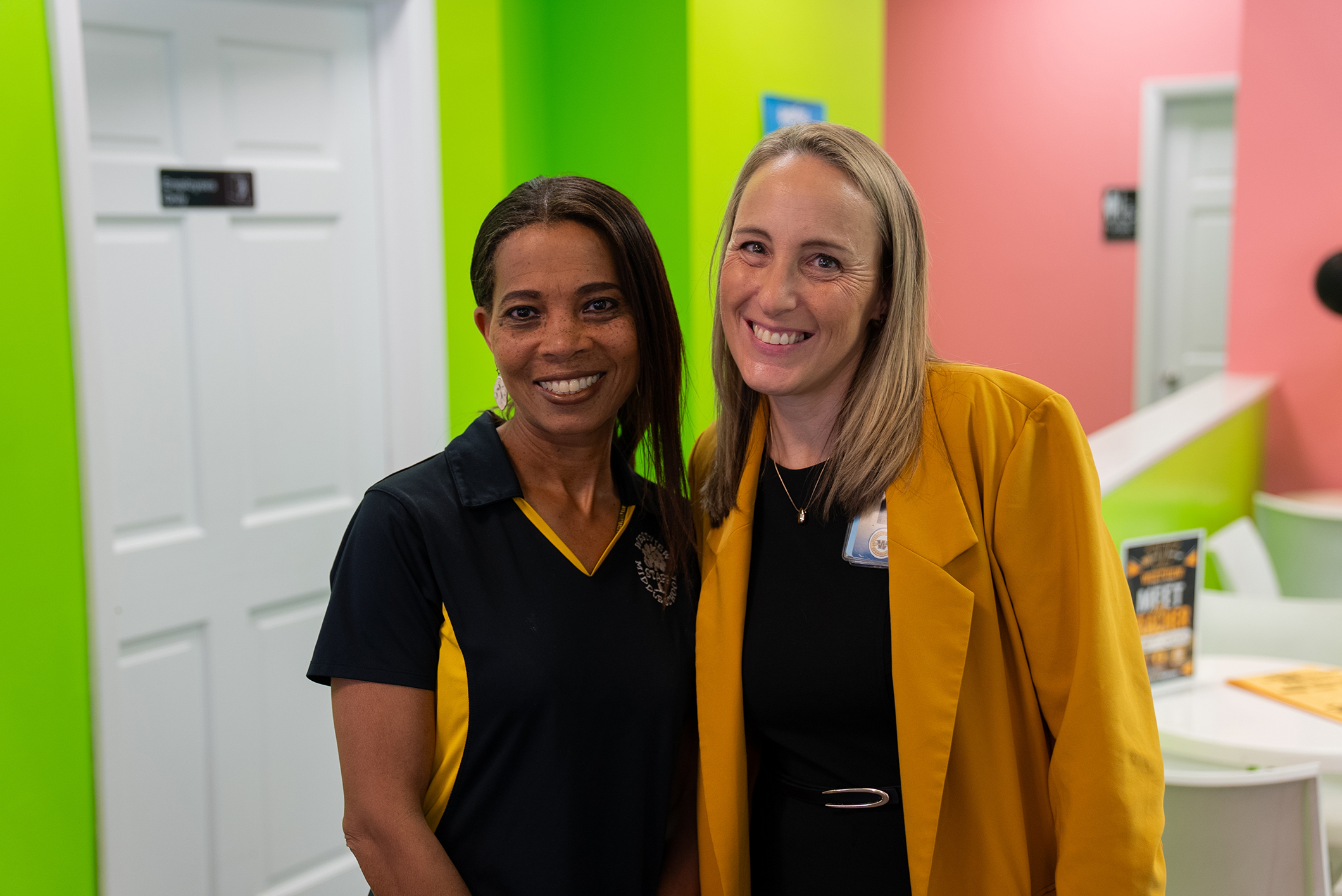 Two women smiling, one in black, the other in yellow, in a brightly colored room with green and pink walls.