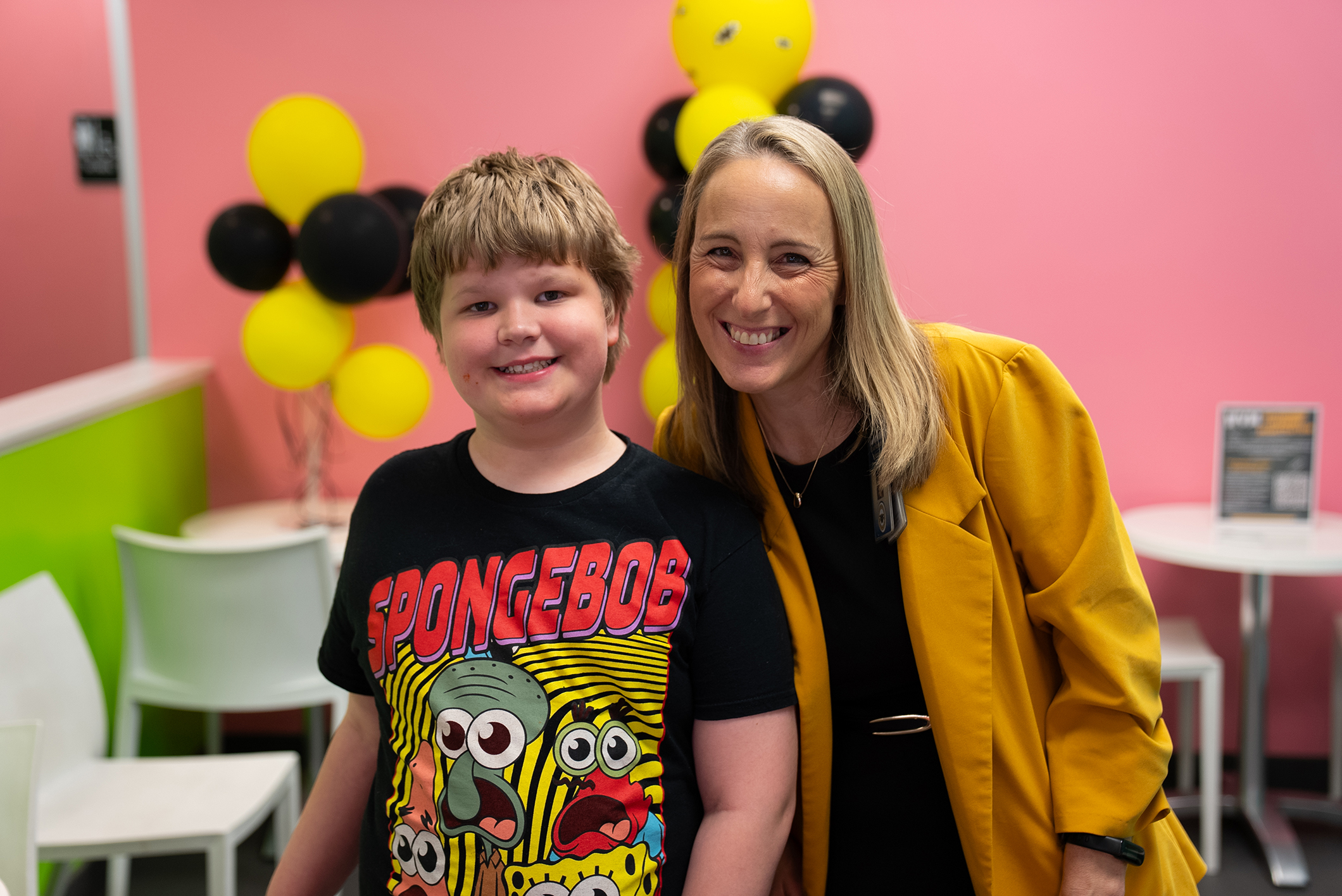A woman with blonde hair and a yellow jacket stands next to a boy in a Spongebob t-shirt.