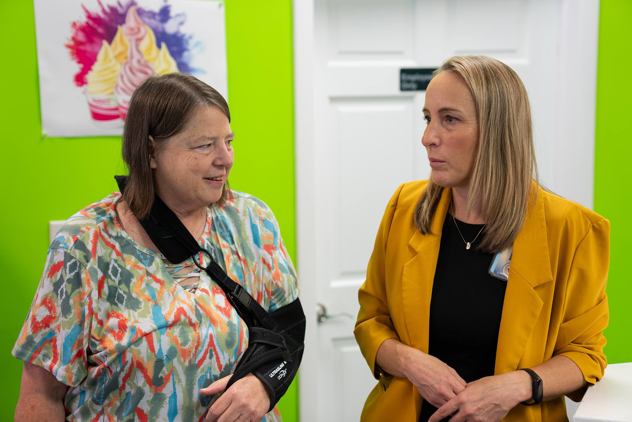 Two women stand in front of a green wall. One wears a colorful top, the other a yellow blazer.
