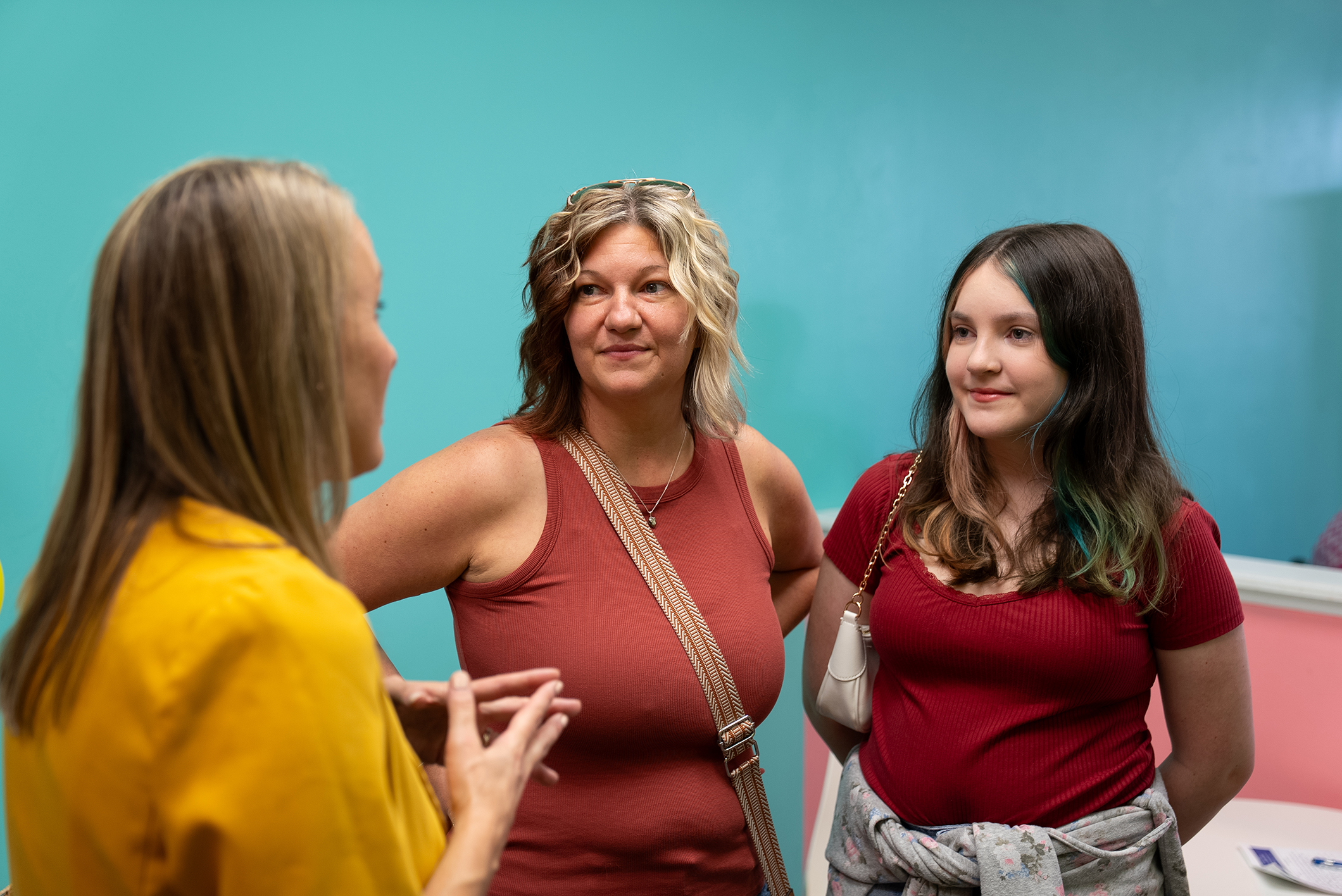 Three women stand in front of a light blue wall; one wears a yellow shirt, another a red shirt, and the third a maroon tank.